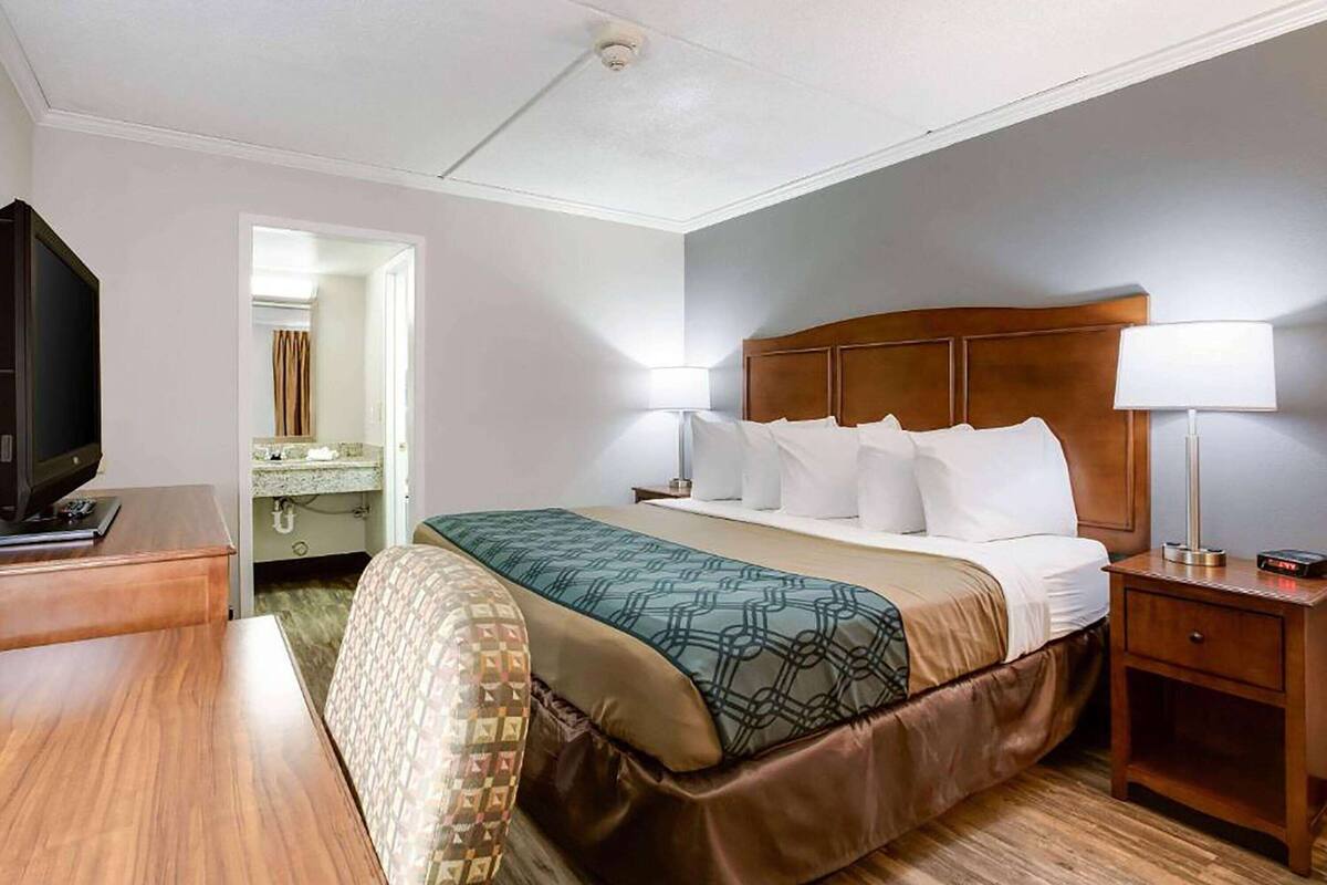 A hotel room with a bed, bedside lamps, a TV, a desk, and a bathroom visible through an open door.