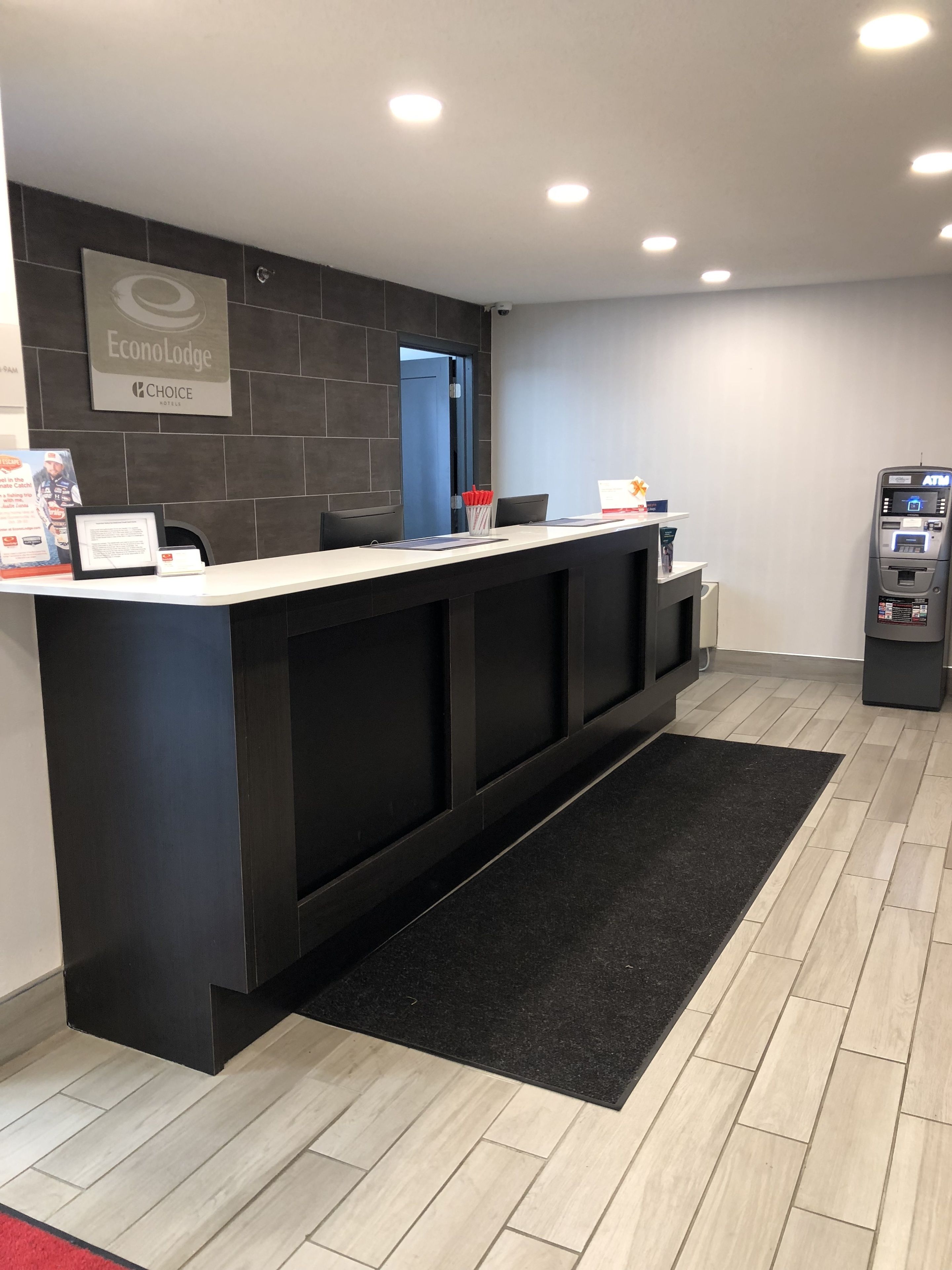 Un comptoir d’accueil avec un revêtement en bois foncé, une moquette noire et un plancher en bois clair. On peut voir une enseigne indiquant « EconoLodge » et « hôtel de qualité ». Un distributeur automatique de billets est visible sur la droite.