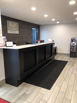 A reception desk with a dark wood finish, a black carpet, and a light wood floor. There is a sign for EconoLodge and a choice hotel. An ATM is visible to the right. - Travelodge by Wyndham Canton/Livonia Area, MI (Canton)