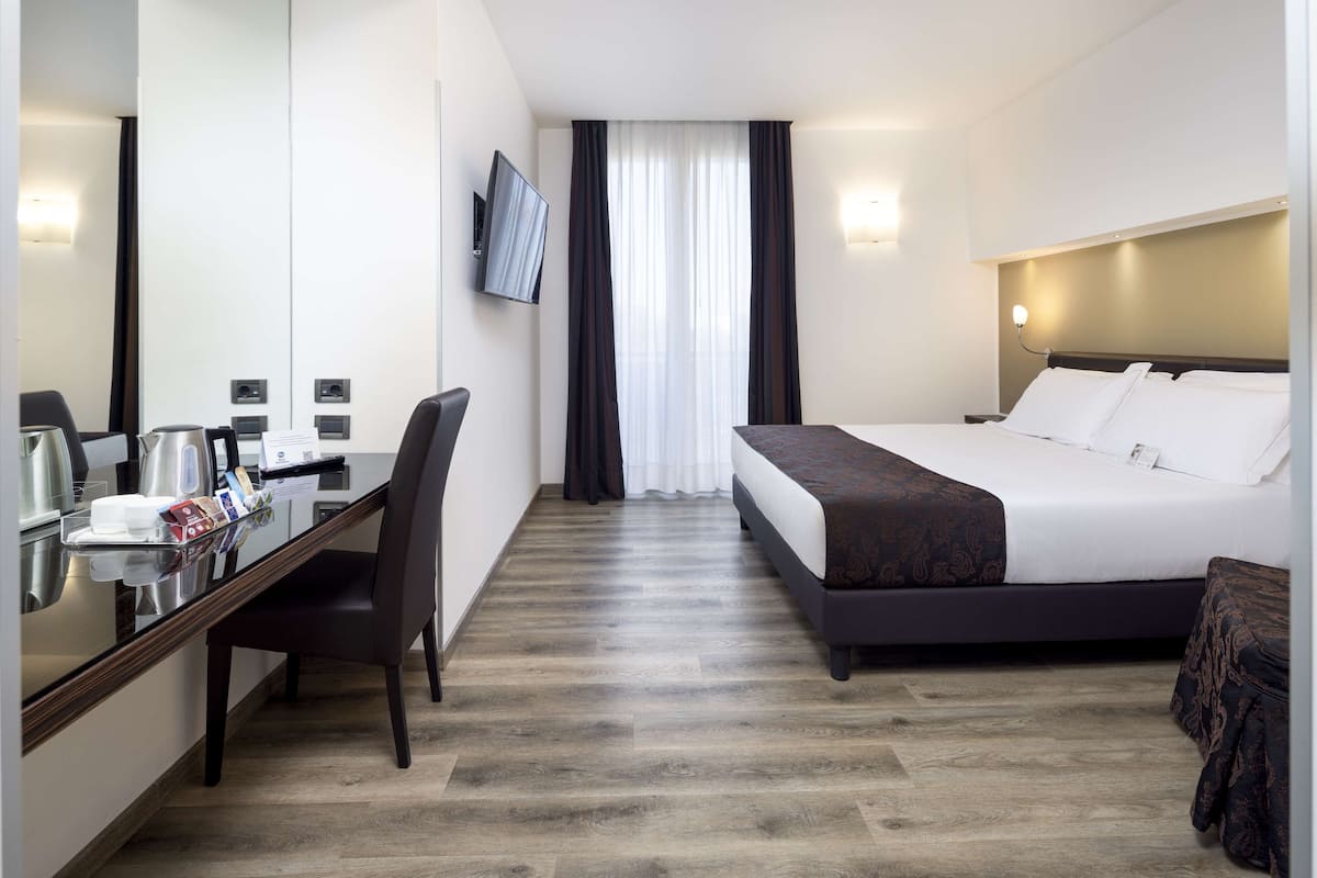 Une chambre d’hôtel comprenant un lit, un bureau avec une chaise, une télévision, une machine à café et une fenêtre avec des rideaux.