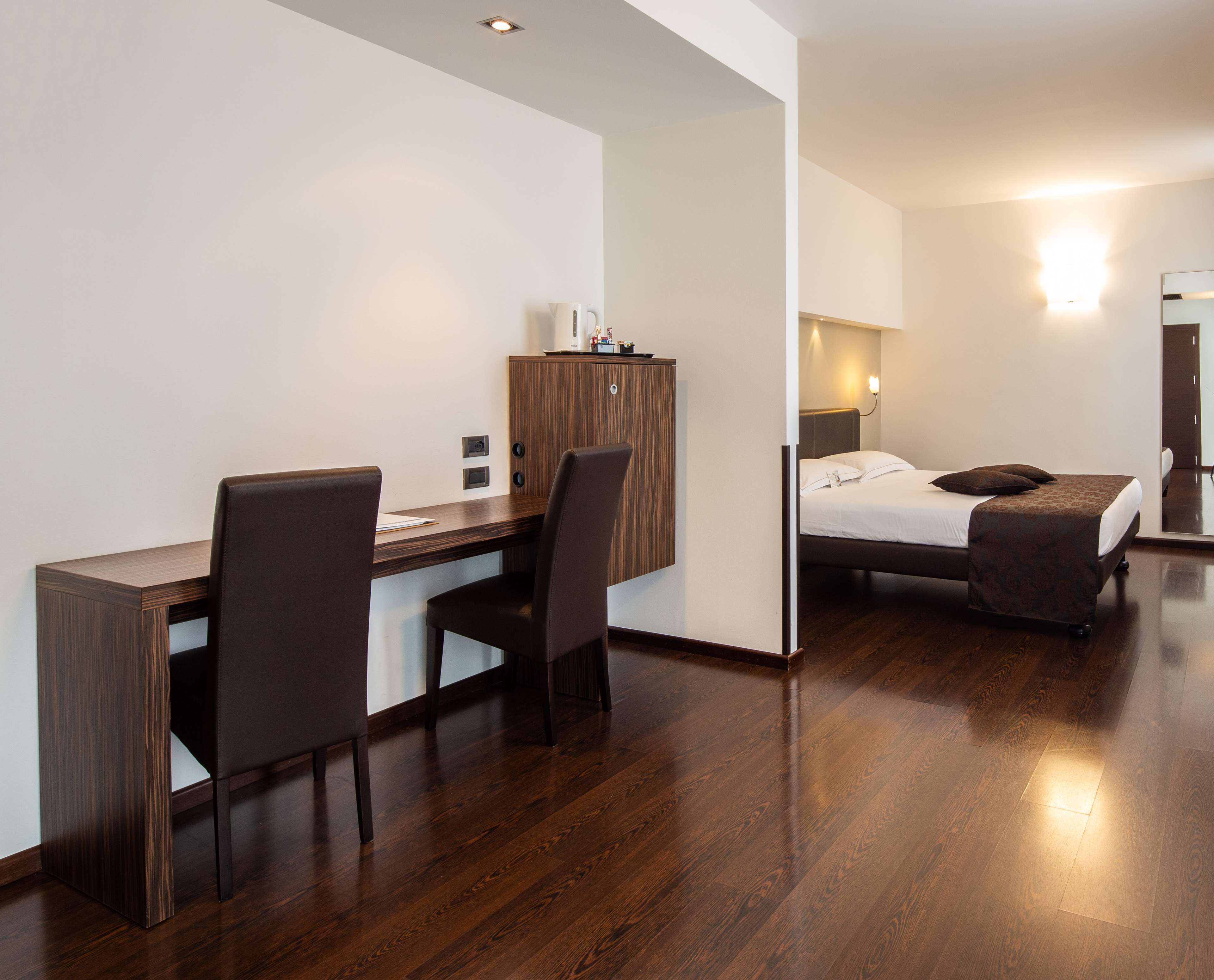 Ein modernes Hotelzimmer mit einem Holztisch, zwei Stühlen, einem Bett mit gemustertem Überwurf und einem Nachttisch mit Lampe.