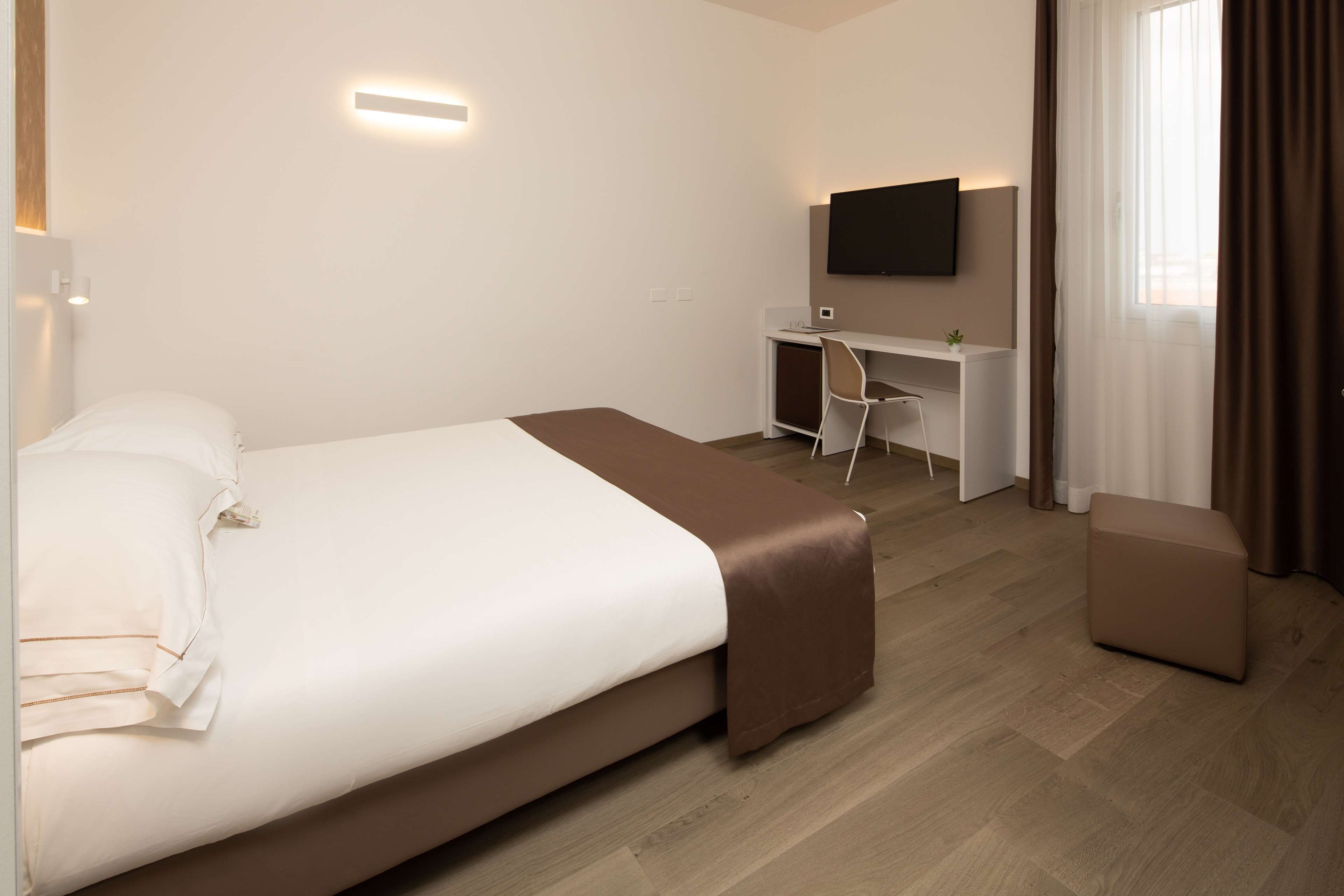 Habitación de hotel con cama, escritorio, silla, televisor y ventana con cortinas.