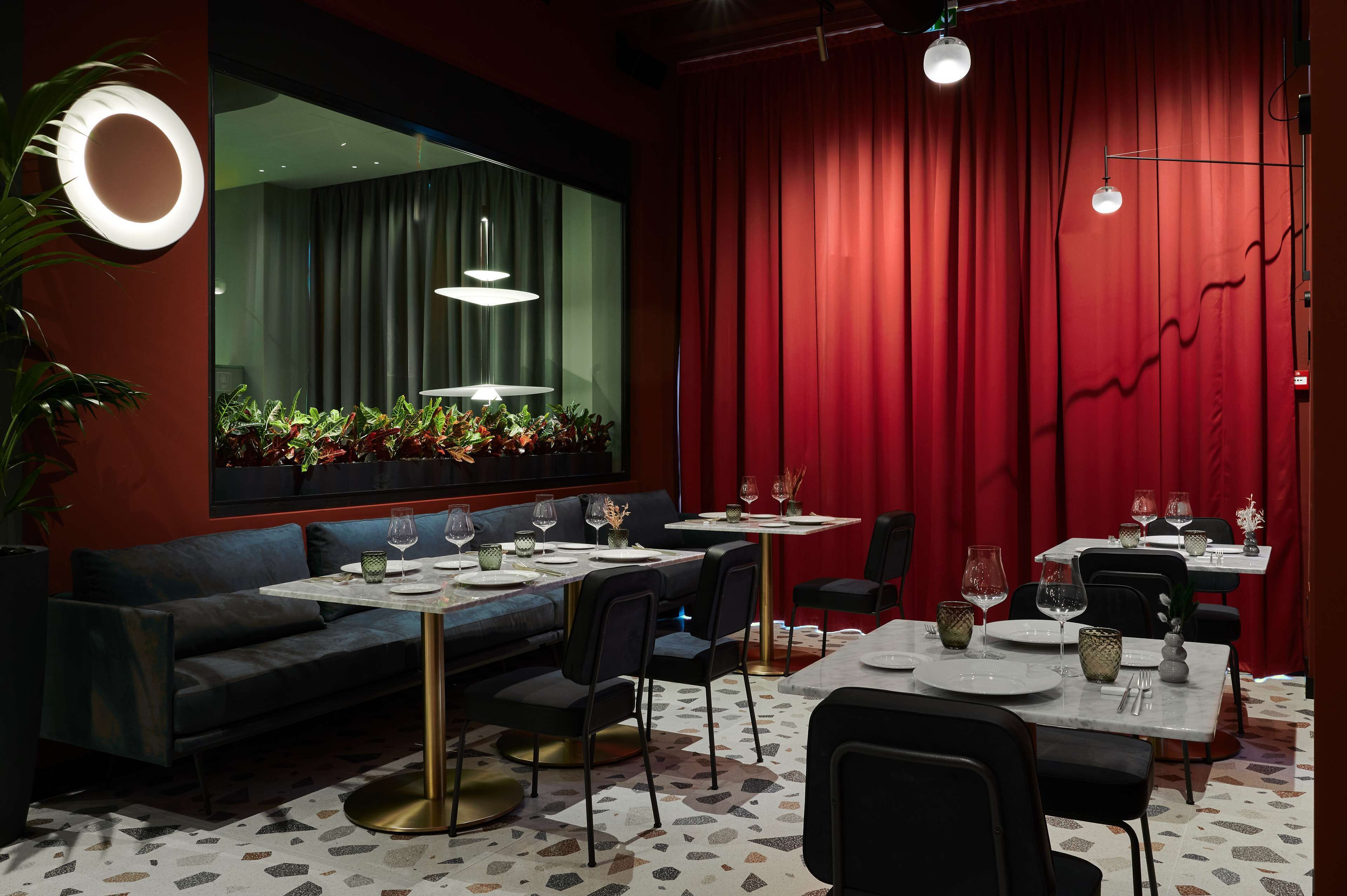 A modern dining area with tables set for two, featuring a red curtain backdrop and ambient lighting.