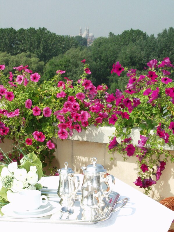 Una mesa dispuesta con cubiertos y teteras, rodeada de flores rosas vibrantes, con vista a un edificio lejano.