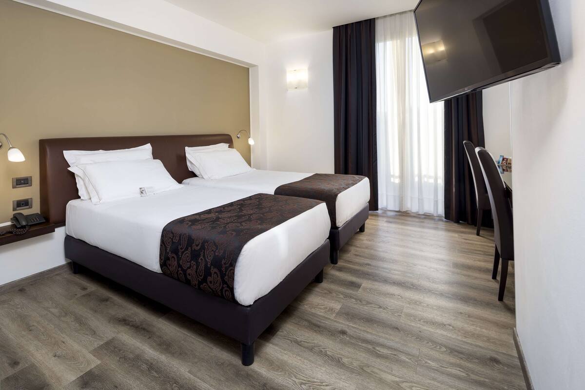 Une chambre d’hôtel avec deux lits, un téléviseur à écran plat, un bureau avec une chaise et une fenêtre avec des rideaux.