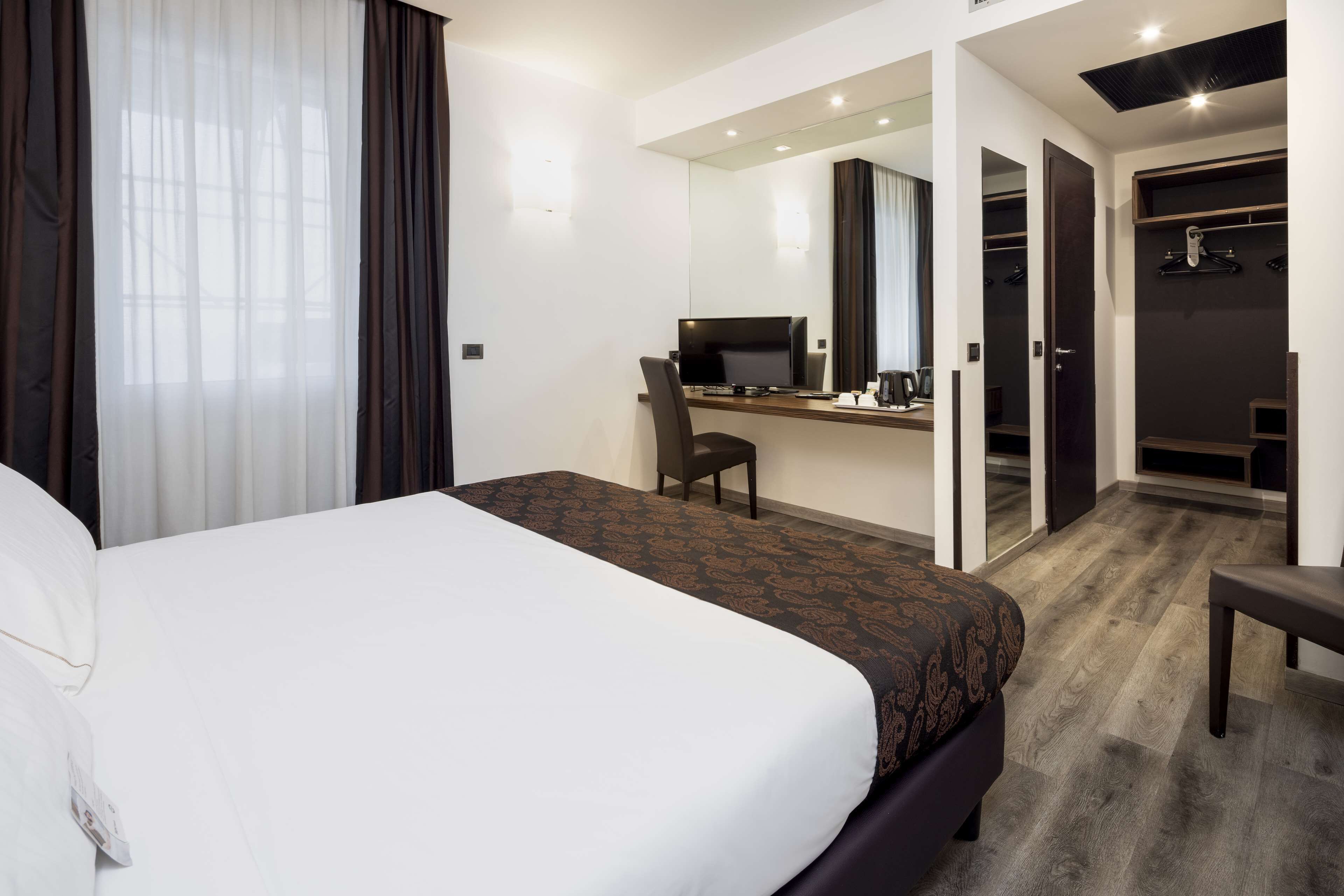 A hotel room with a large bed, a desk with a computer, a chair, a mirror, and a walk-in closet.