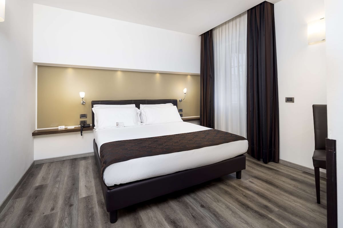 Une chambre d’hôtel moderne avec un grand lit, deux tables de chevet, une chaise et une grande fenêtre avec des rideaux.