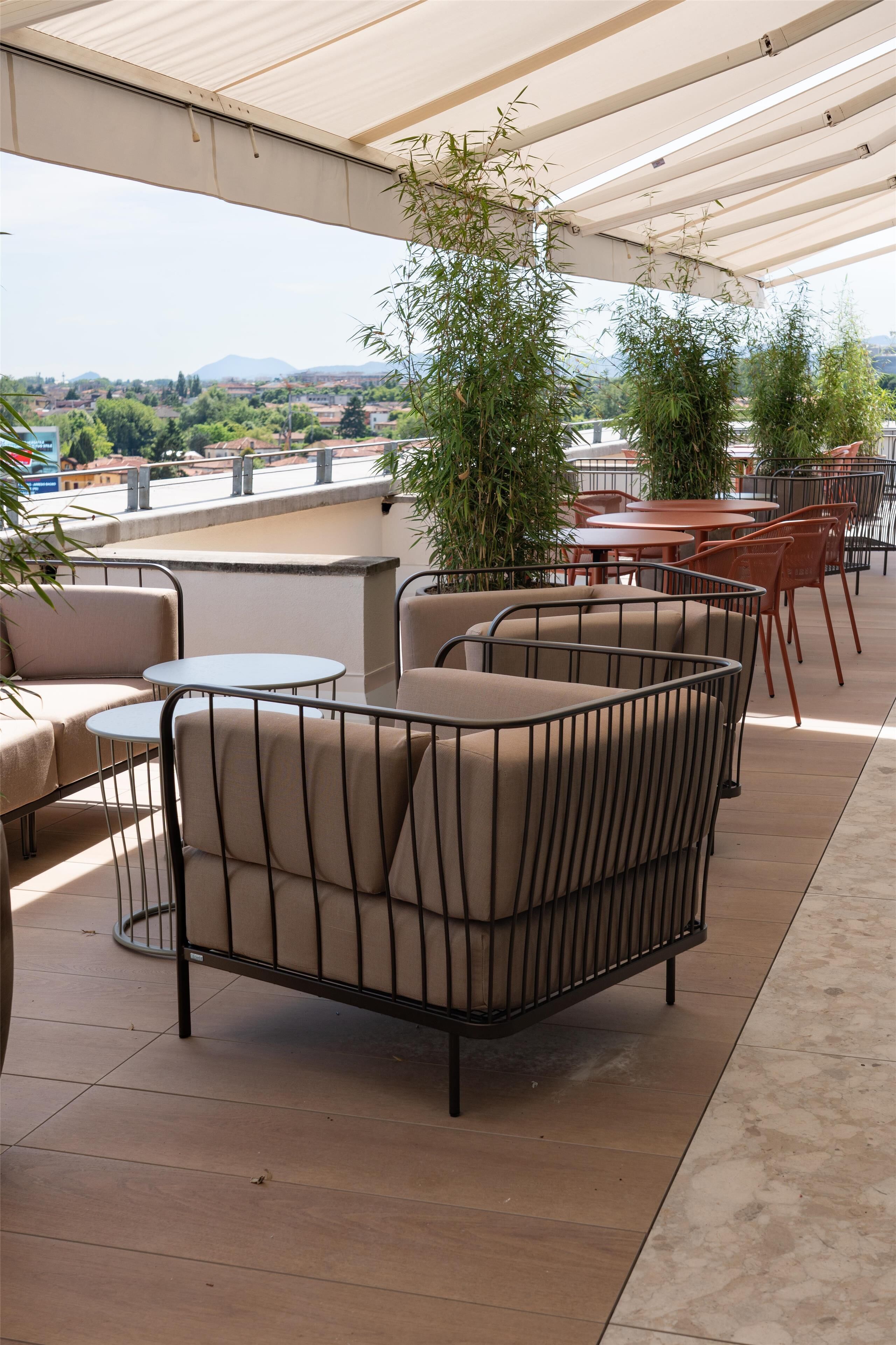 Zona relax all'aperto con sedie e tavoli imbottiti, circondata da un portico coperto e vista sulla città.