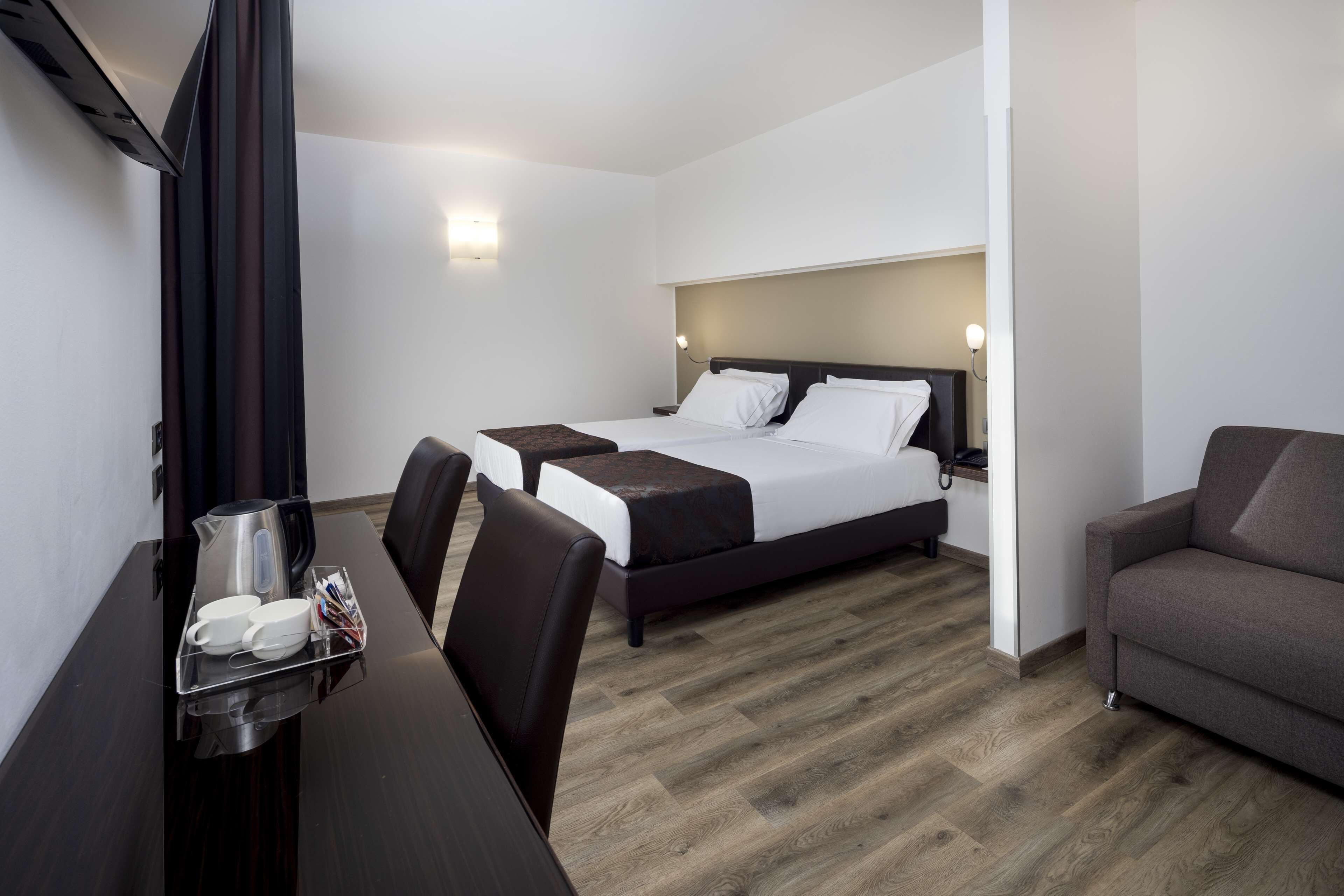 A hotel room with a bed, a desk with a tray of cups and a kettle, a sofa, and a wooden floor.