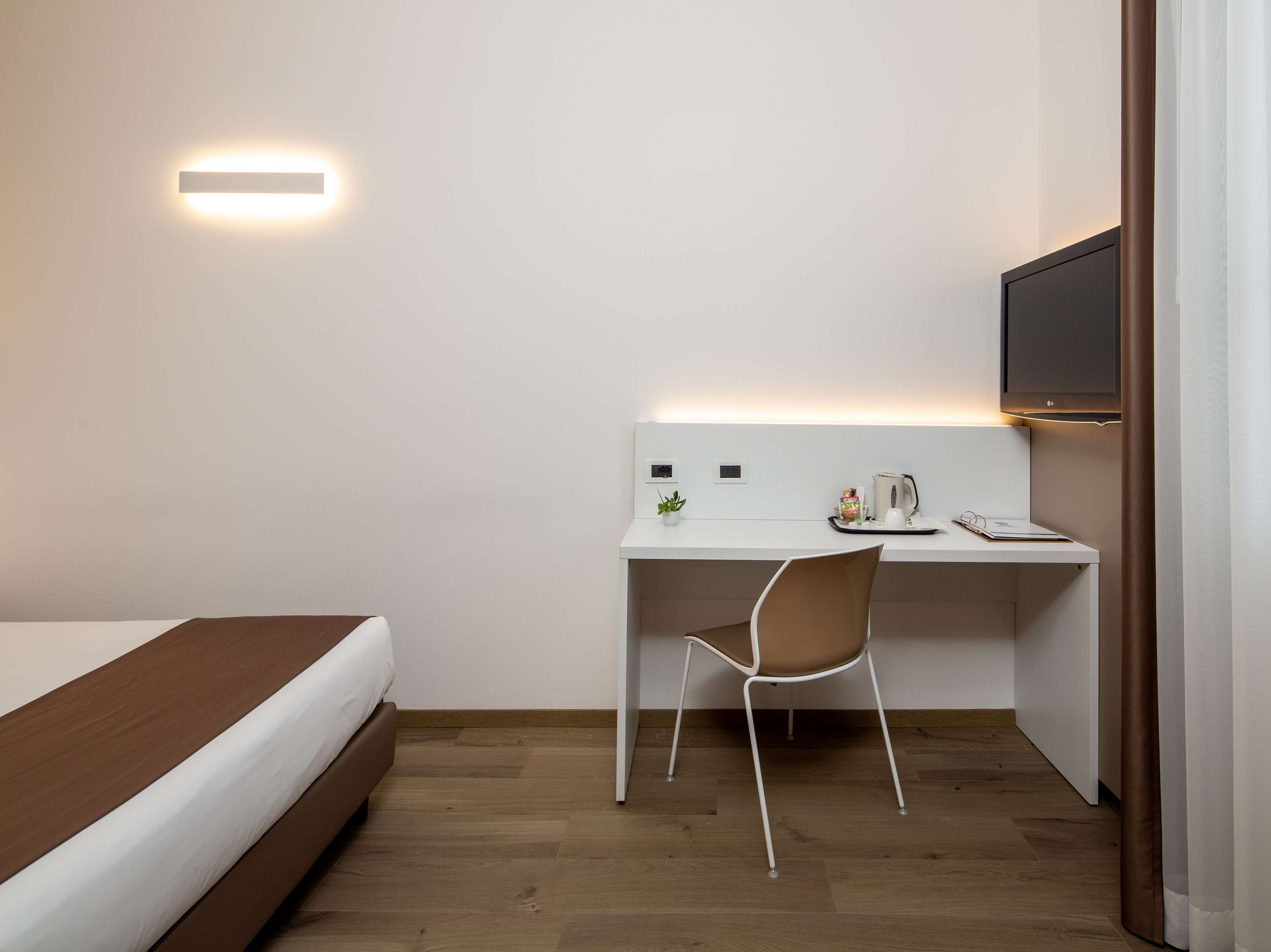 Quarto de hotel com cama, escrivaninha com cadeira, televisão de tela plana e luminária na parede.