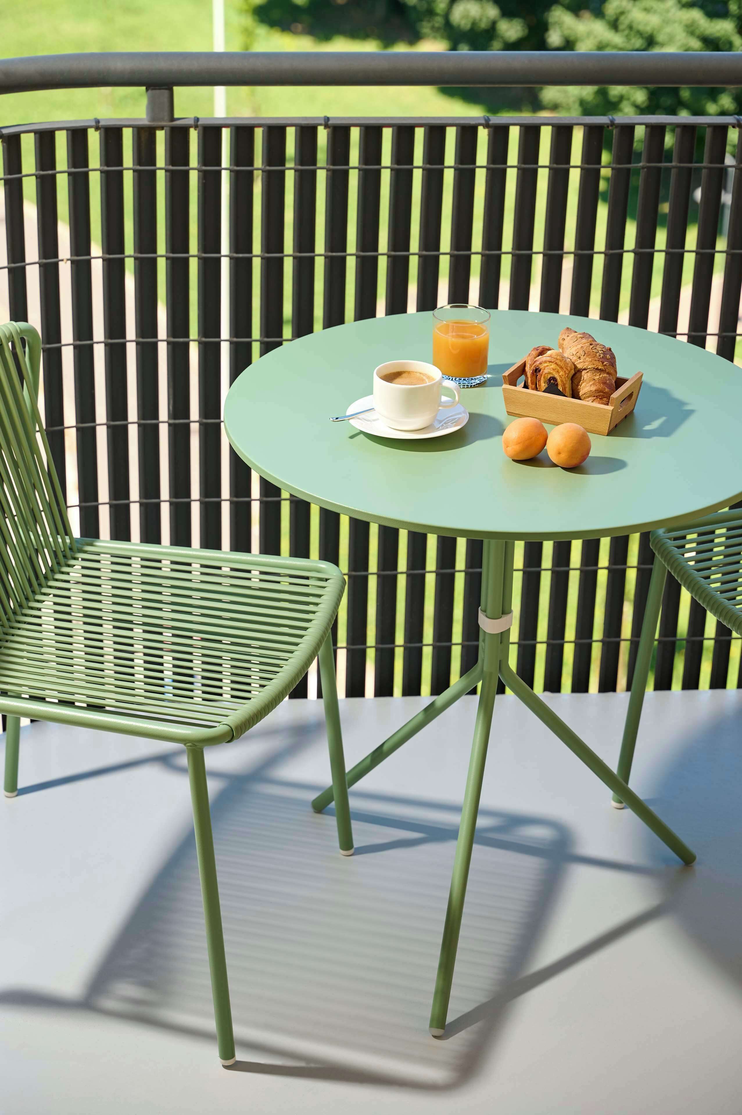 A green metal chair and table with a cup of coffee, croissants, and oranges on a balcony.