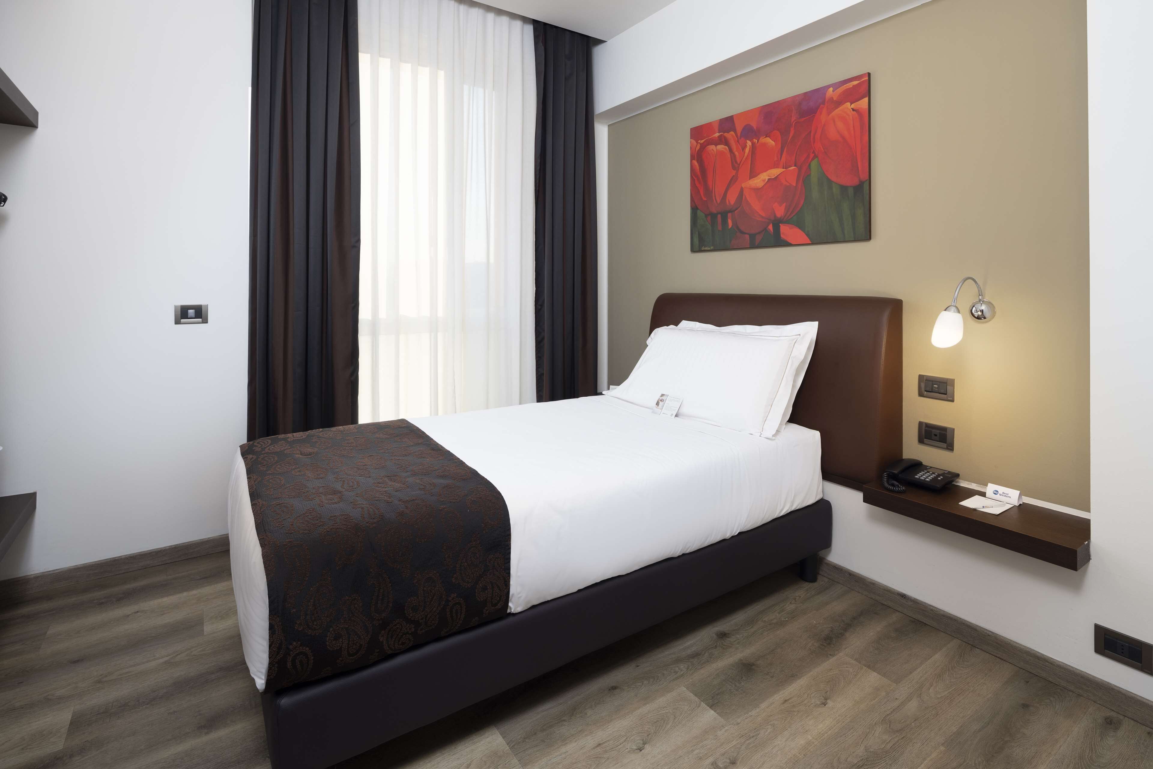 A hotel room with a bed, a nightstand, a painting, and a window with curtains.