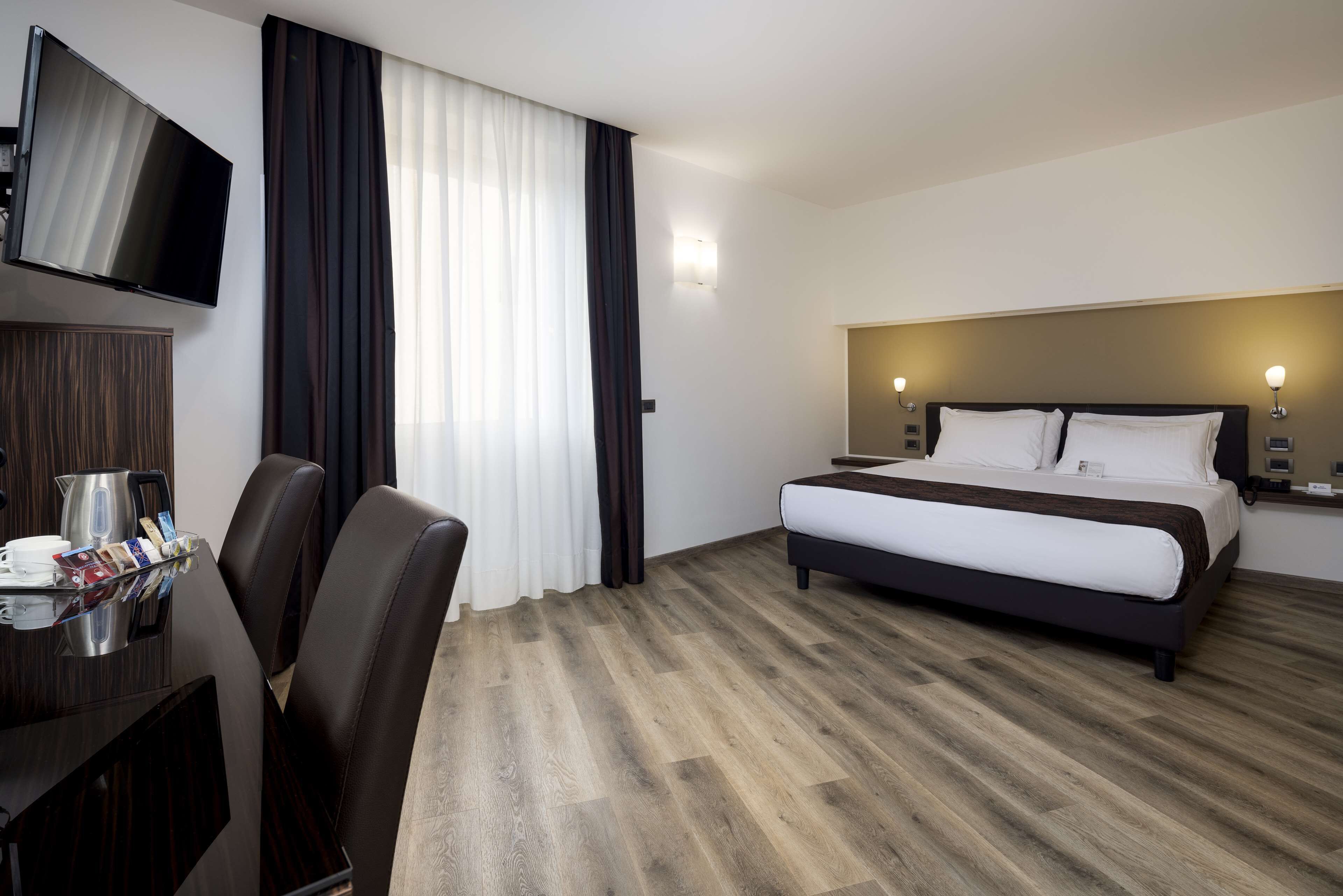 A hotel room with a bed, a TV mounted on the wall, a desk with a coffee maker, and a chair.