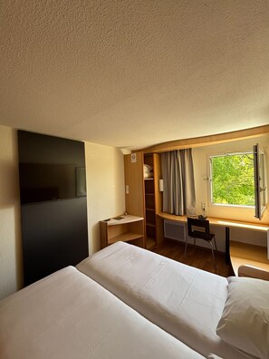 Une chambre d’hôtel avec un lit, un bureau, une chaise, une télévision et une fenêtre avec des rideaux.