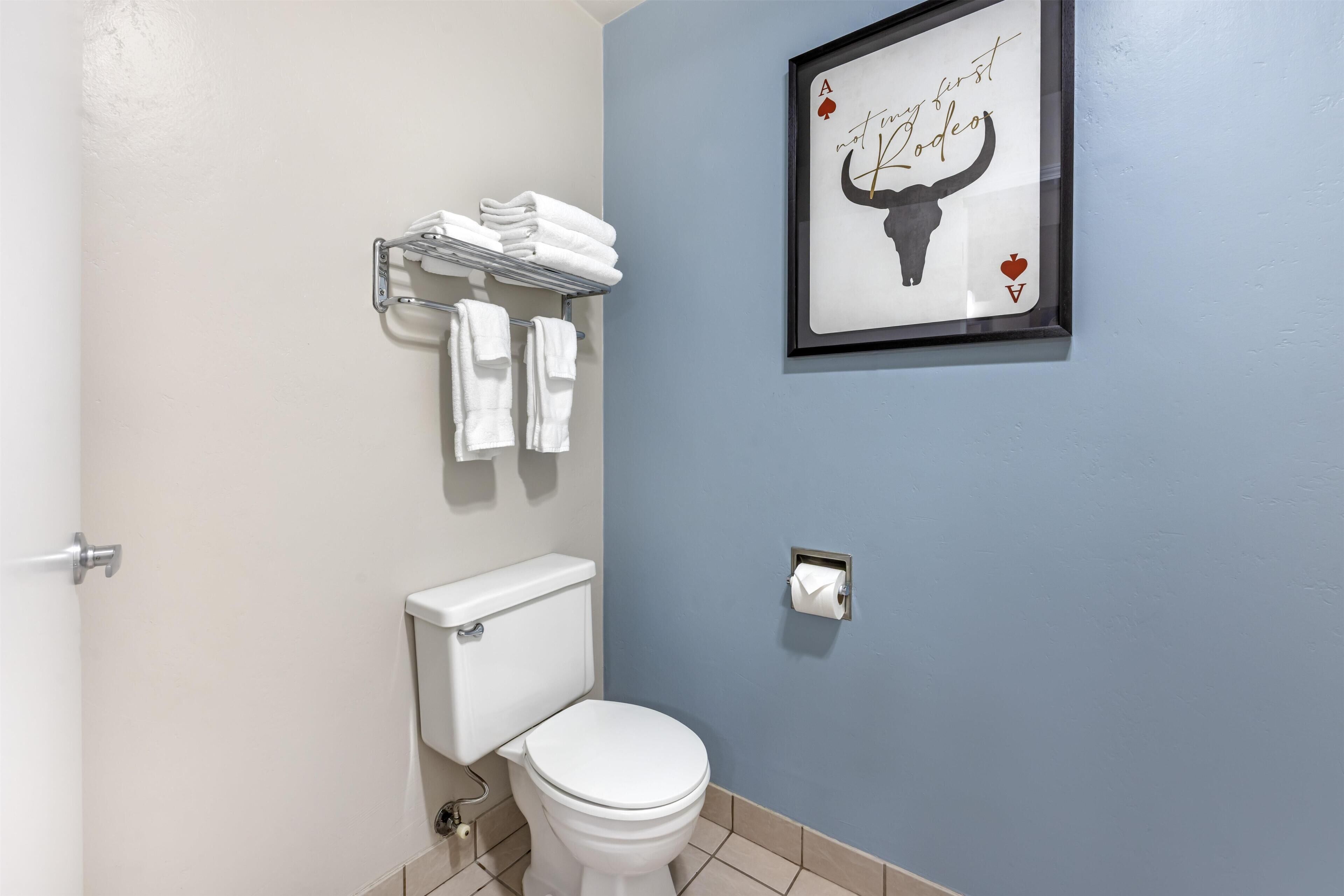 A bathroom with a toilet, a towel rack with folded towels, and a framed picture on the wall.