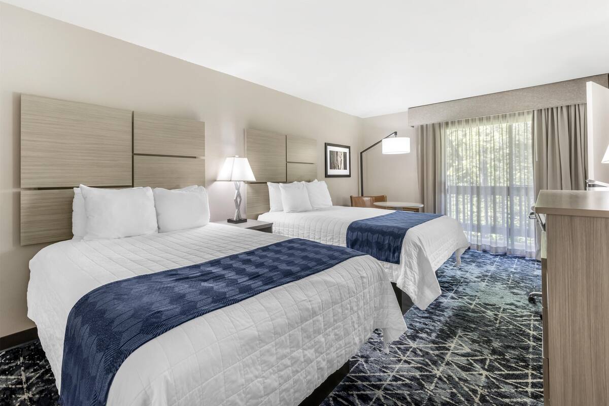 A hotel room with two beds, a desk, a chair, a lamp, and a large window with curtains.