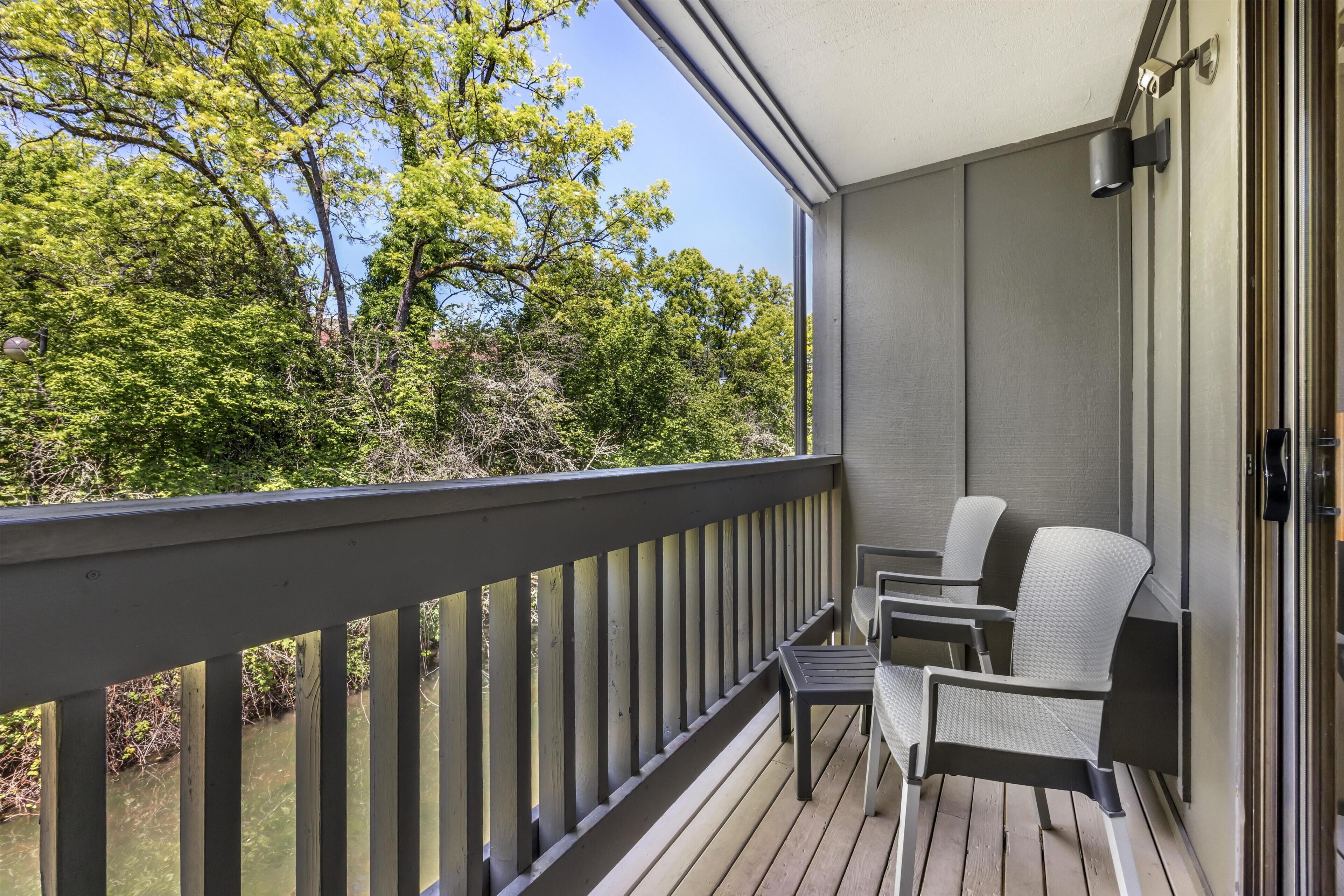 A balcony with a view of trees and a body of water, equipped with two chairs and a small table.