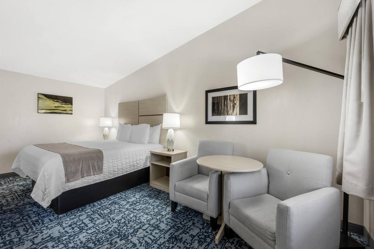 A hotel room with a bed, two bedside tables, a chair, a small round table, and a large window.