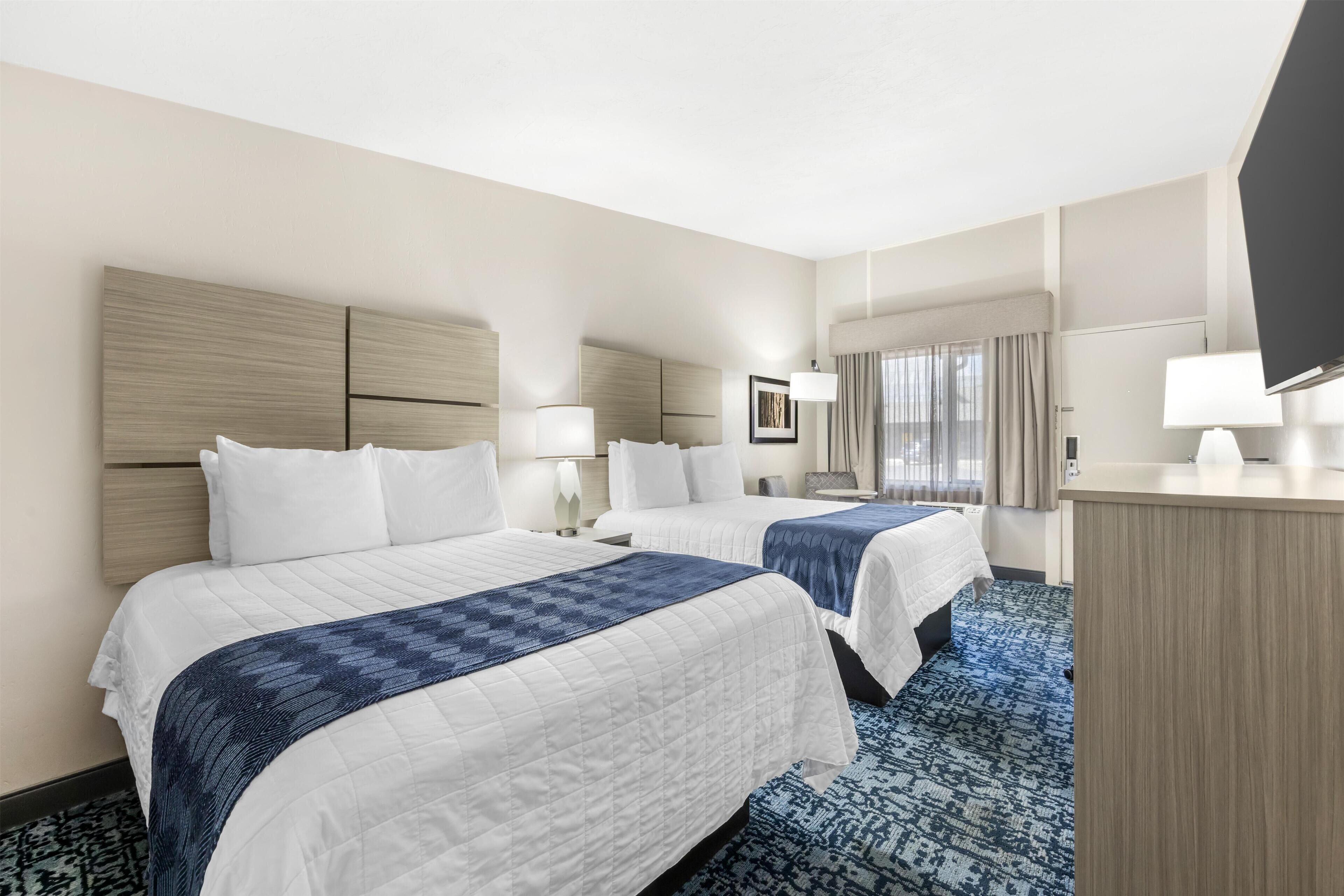 Habitación de hotel con dos camas, un escritorio, un televisor y un ventanal con cortinas.