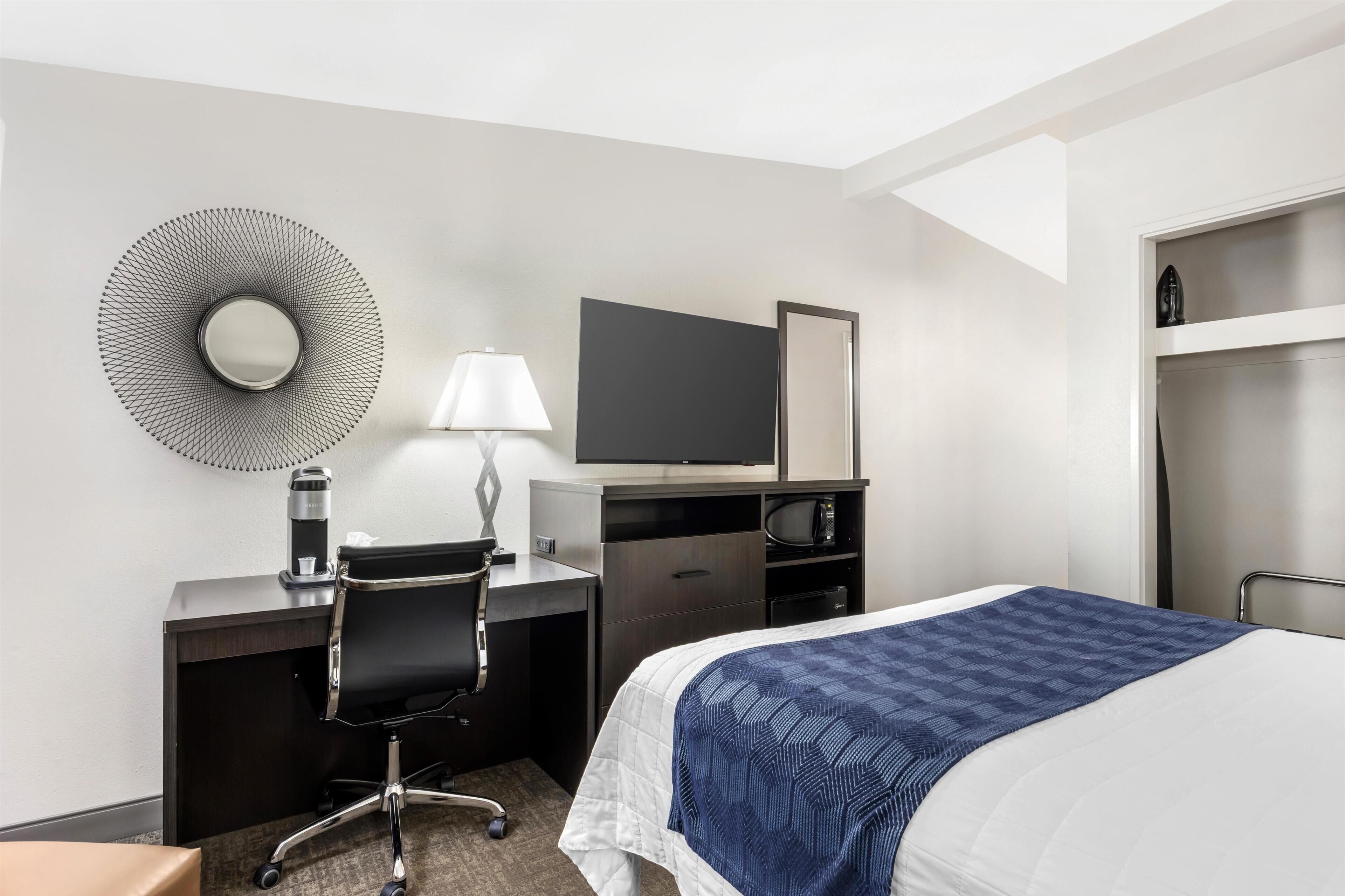 A hotel room with a bed, a desk with a chair, a television, a mirror, and a wardrobe.
