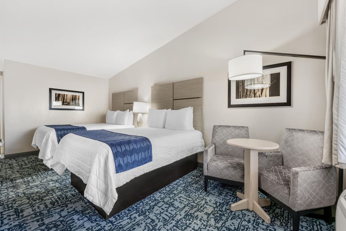 A hotel room with a large bed, a small round table, two armchairs, a lamp, and framed pictures on the walls.