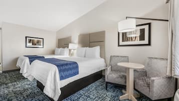 A hotel room with a large bed, a small round table, two armchairs, a lamp, and framed pictures on the walls.