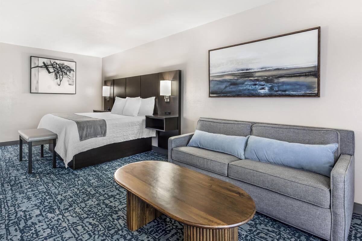 A hotel room with a bed, a sofa, a coffee table, a wall-mounted TV, and artwork.