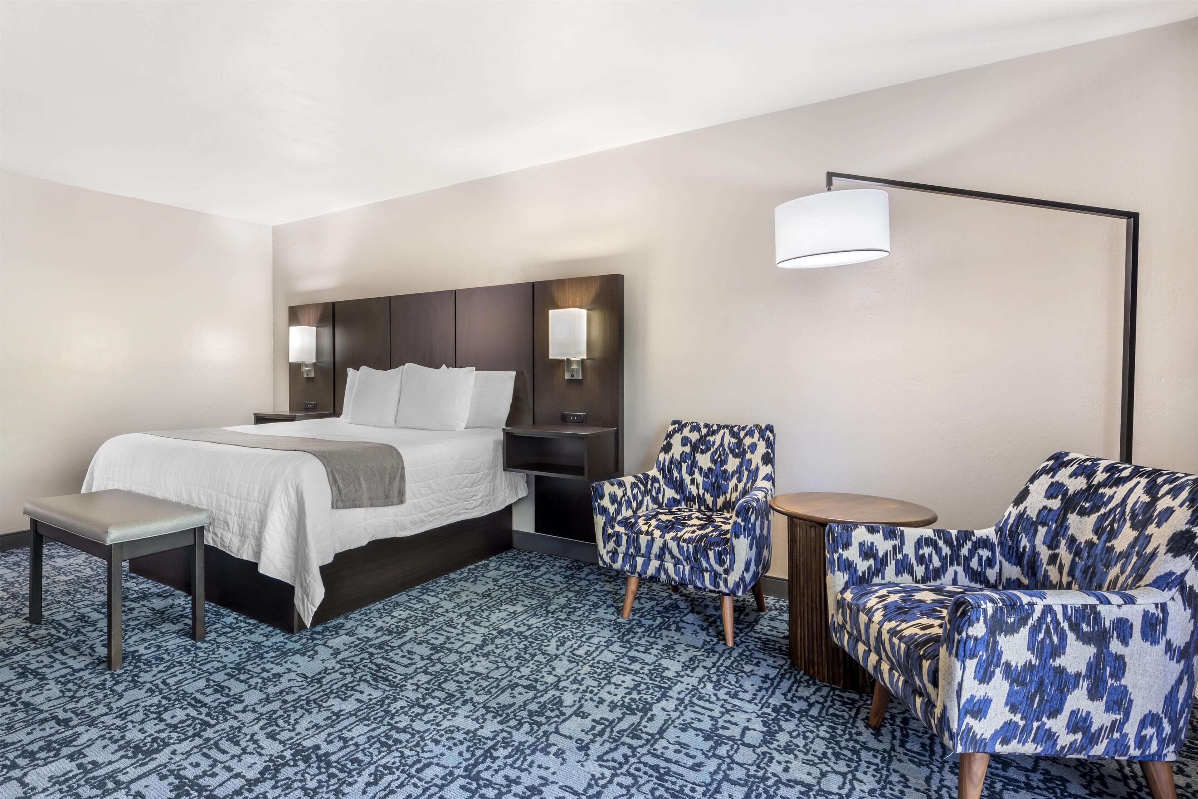 A hotel room with a bed, two armchairs, a small table, a bedside lamp, and a patterned carpet.