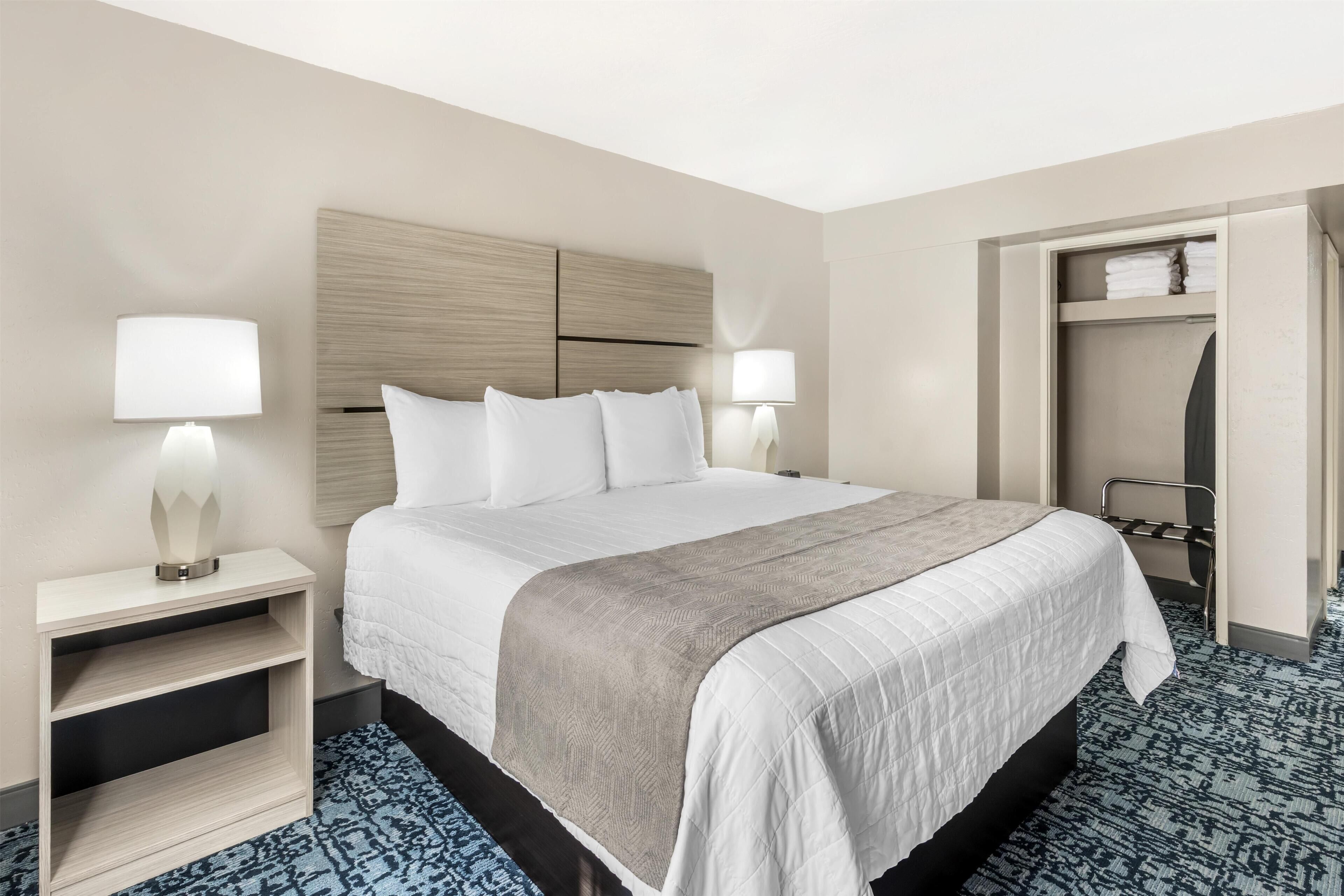 A hotel room with a bed, bedside lamps, a nightstand, a closet, and a patterned carpet.