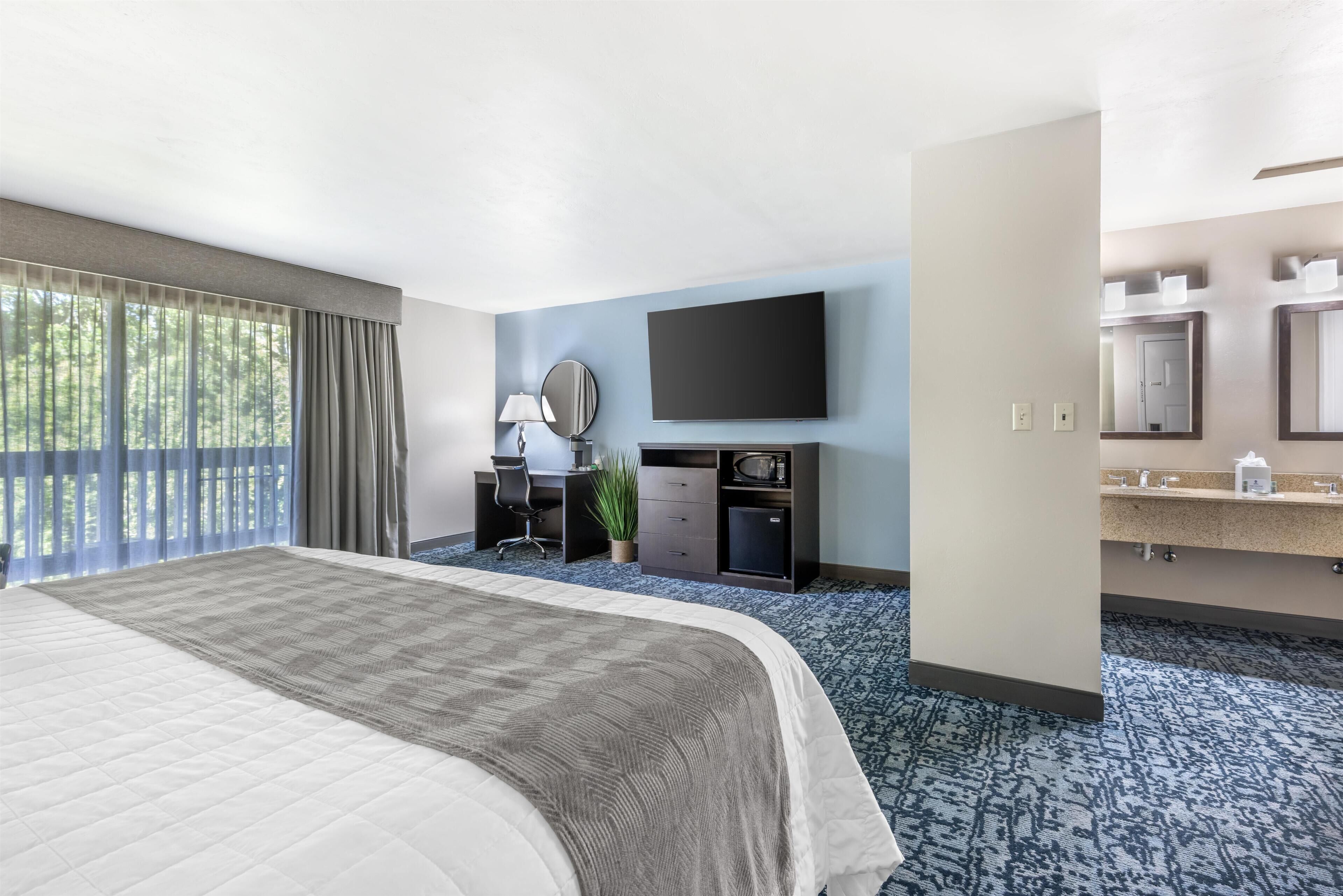 A hotel room with a large bed, a desk with a chair, a flat-screen TV, a mirror, and a bathroom with a sink and mirror.