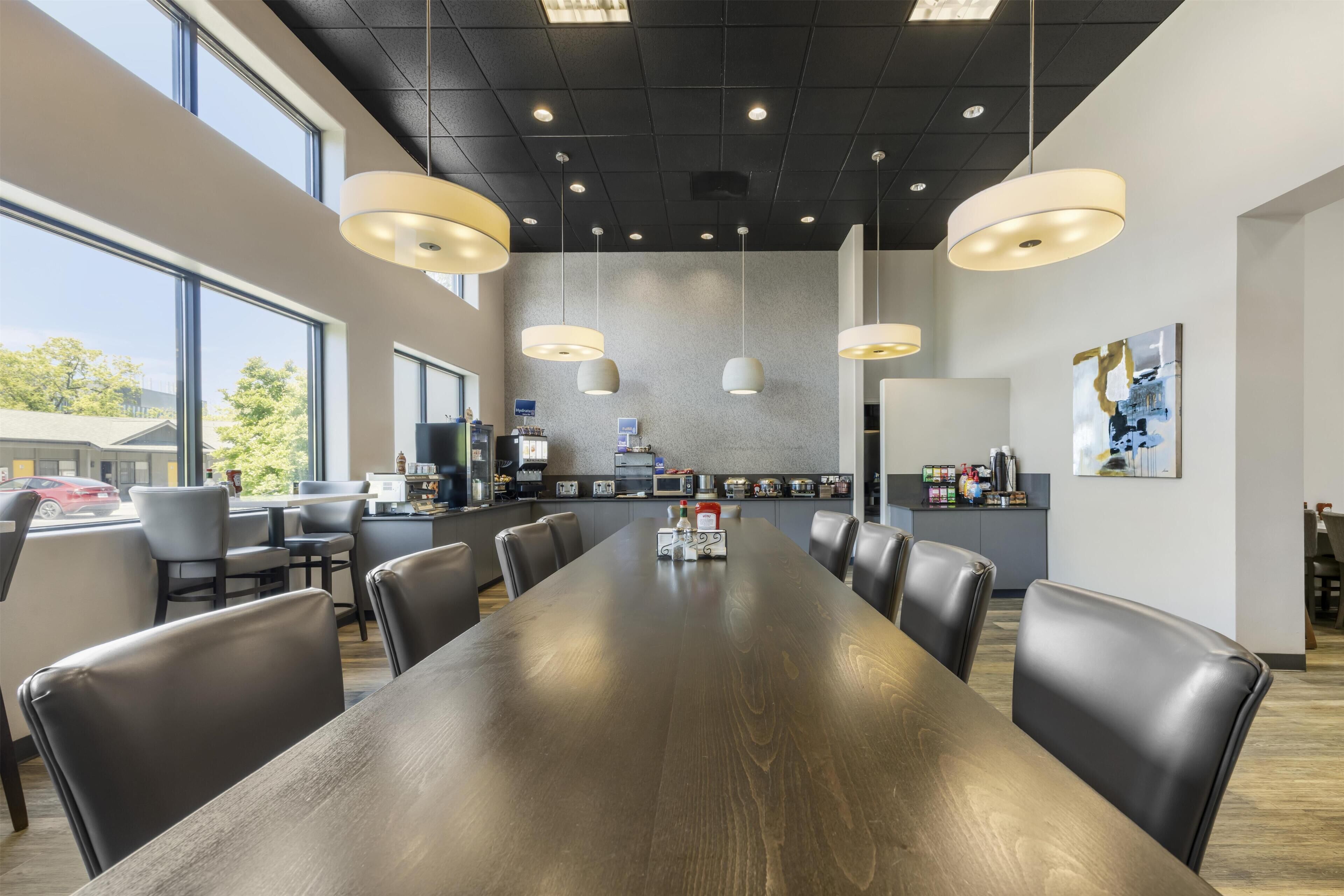 A conference room with a long table, multiple chairs, pendant lights, a kitchen area, and large windows.