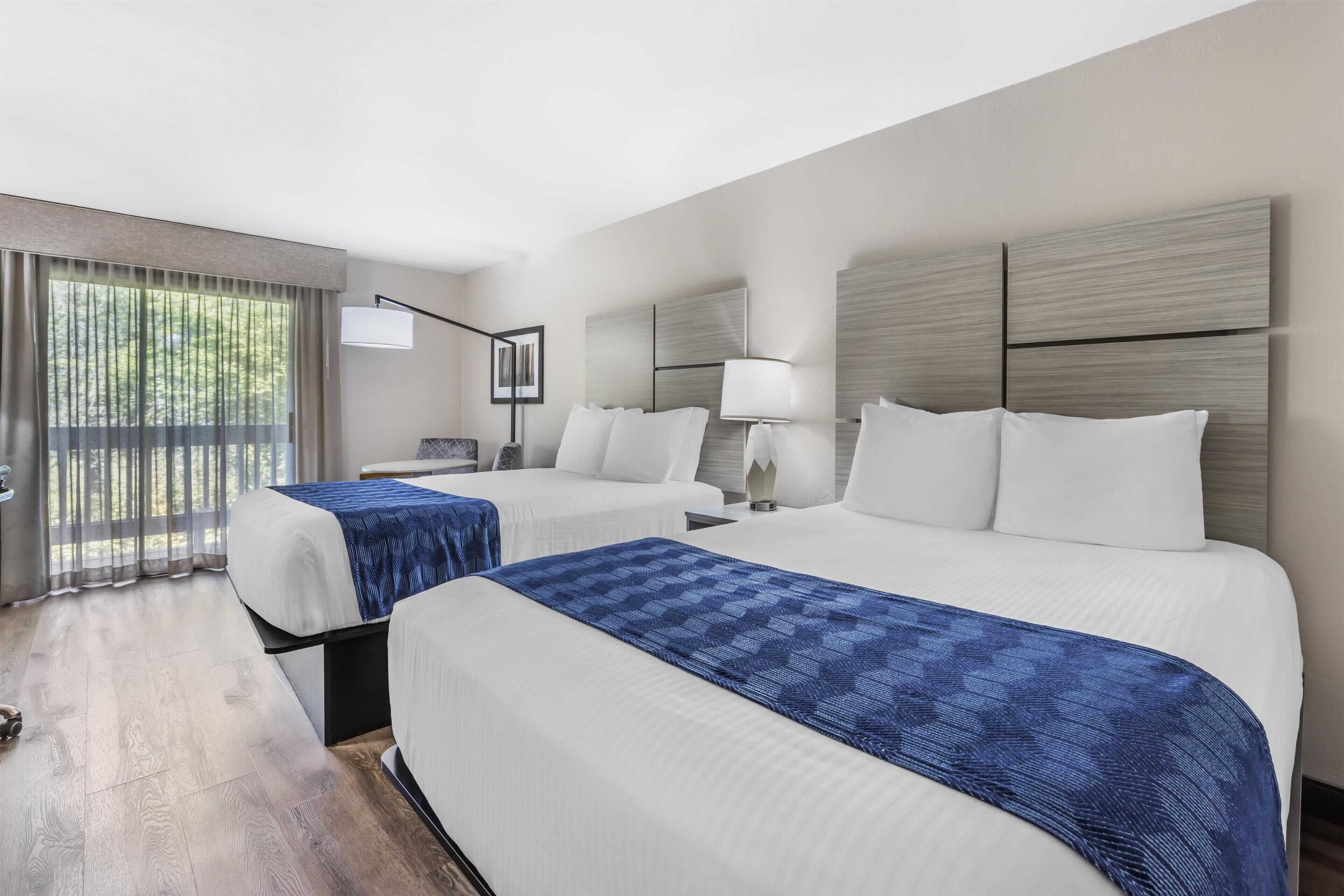 Habitación de hotel con dos camas, un ventanal con cortinas, una mesita de noche con lámpara y un cabecero con panel de luz.