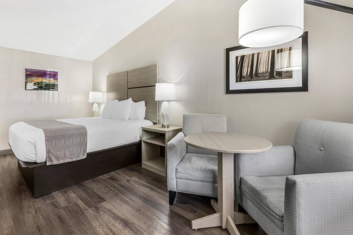 A hotel room with a bed, two bedside tables with lamps, a small round table, a grey armchair, and a framed picture on the wall.