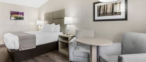A hotel room with a bed, two bedside tables with lamps, a small round table, a grey armchair, and a framed picture on the wall.
