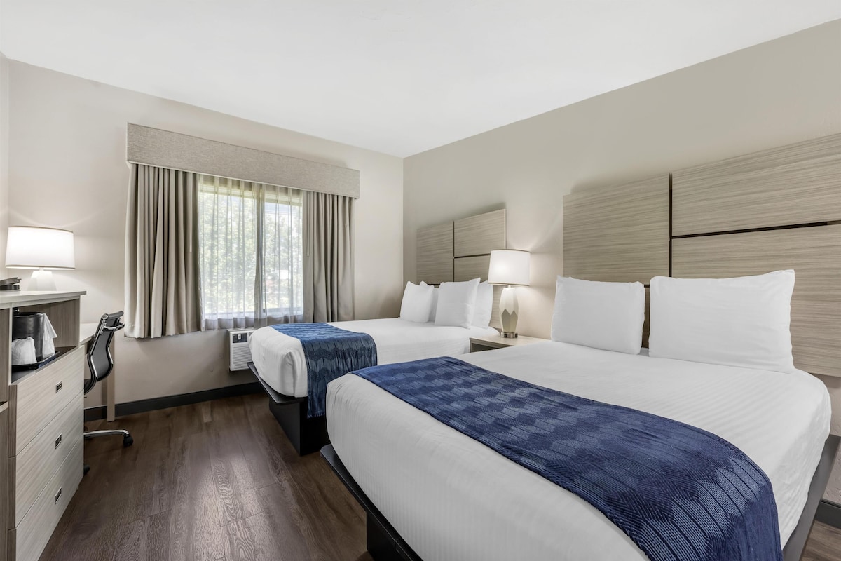A hotel room with two beds, a desk, a chair, a lamp, and a window with curtains.
