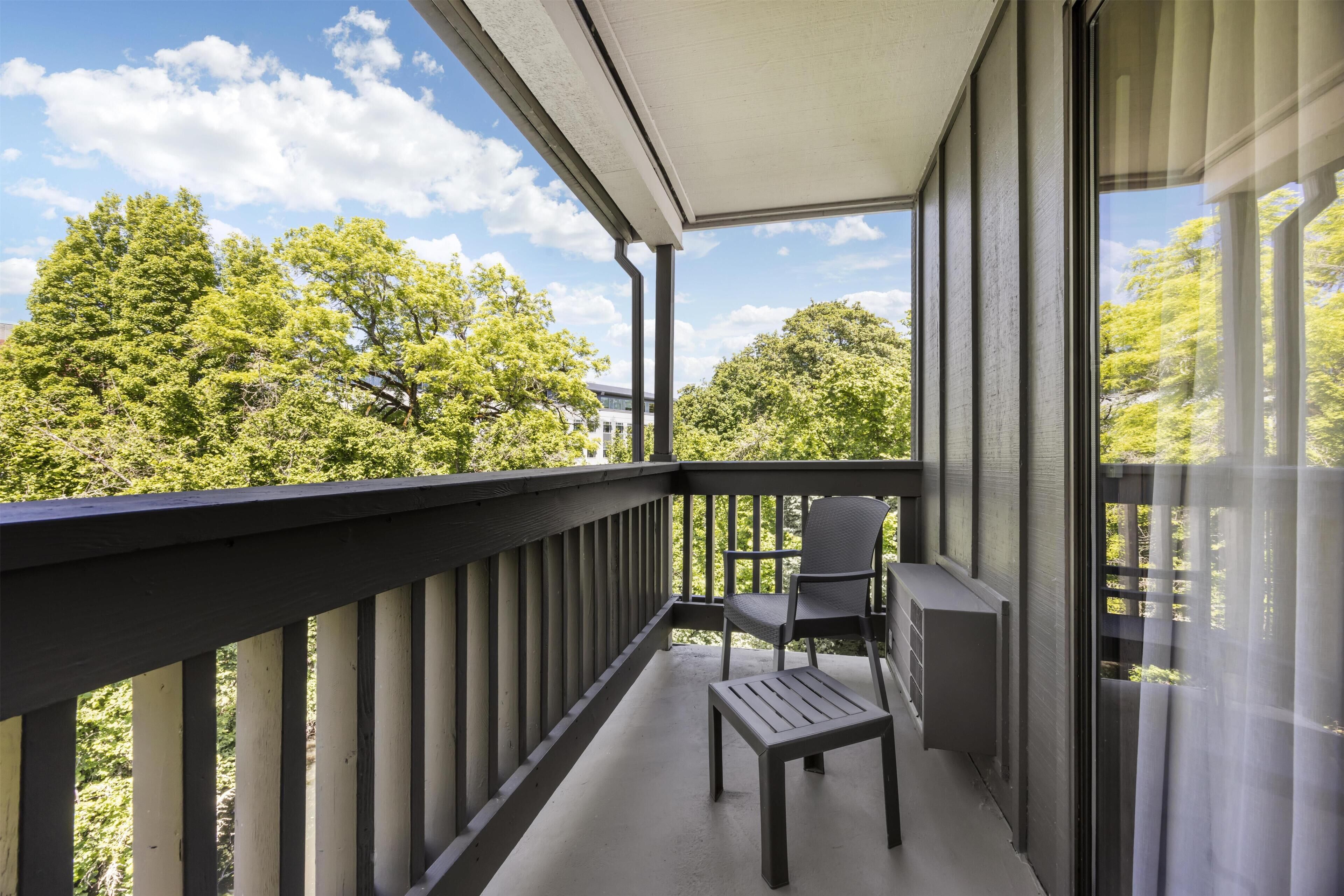 Un balcone con una sedia e un tavolo, affaccia su un paesaggio verde con alberi e un edificio in lontananza.