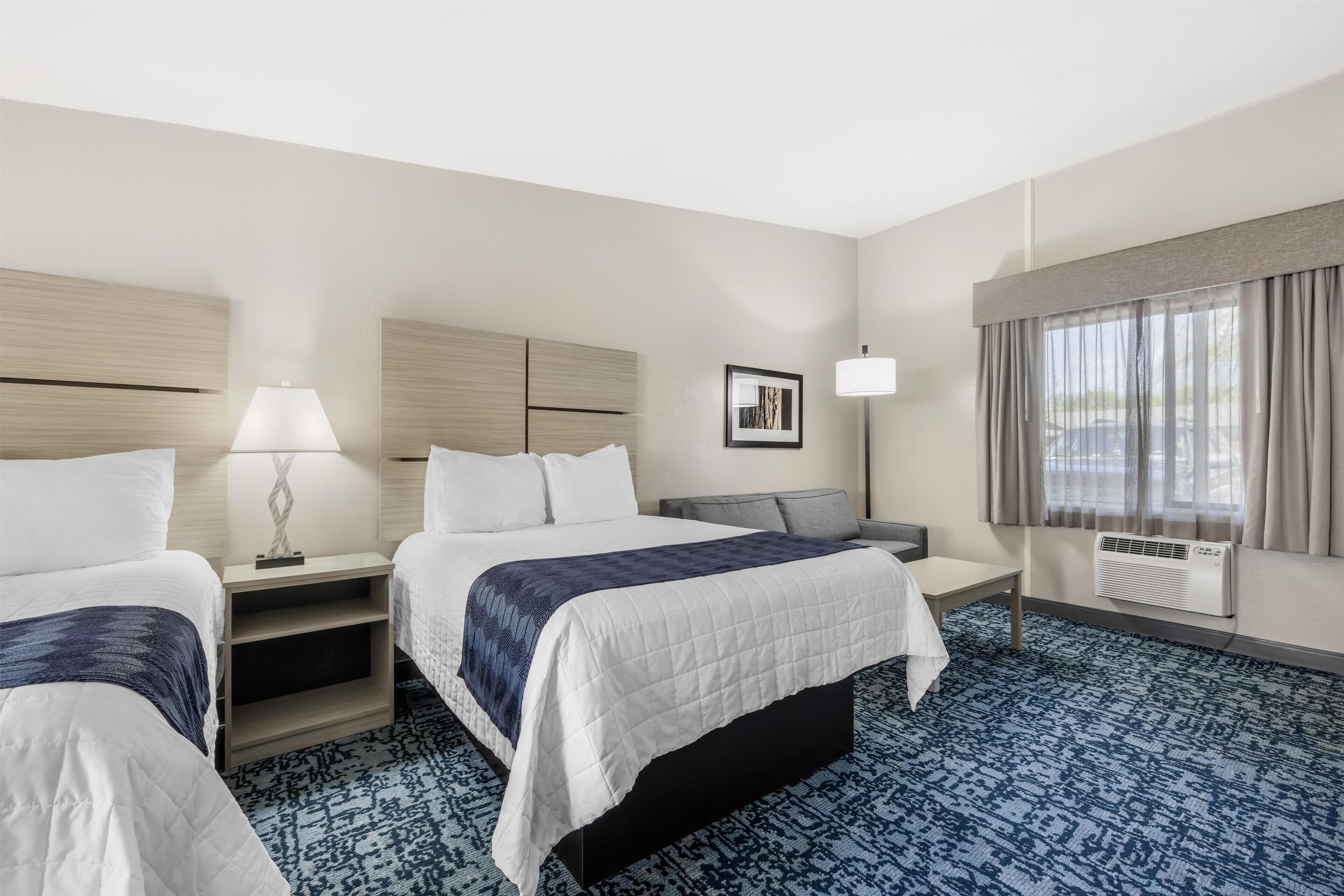 A hotel room with two beds, a desk, a chair, a lamp, a picture on the wall, and a large window with curtains.
