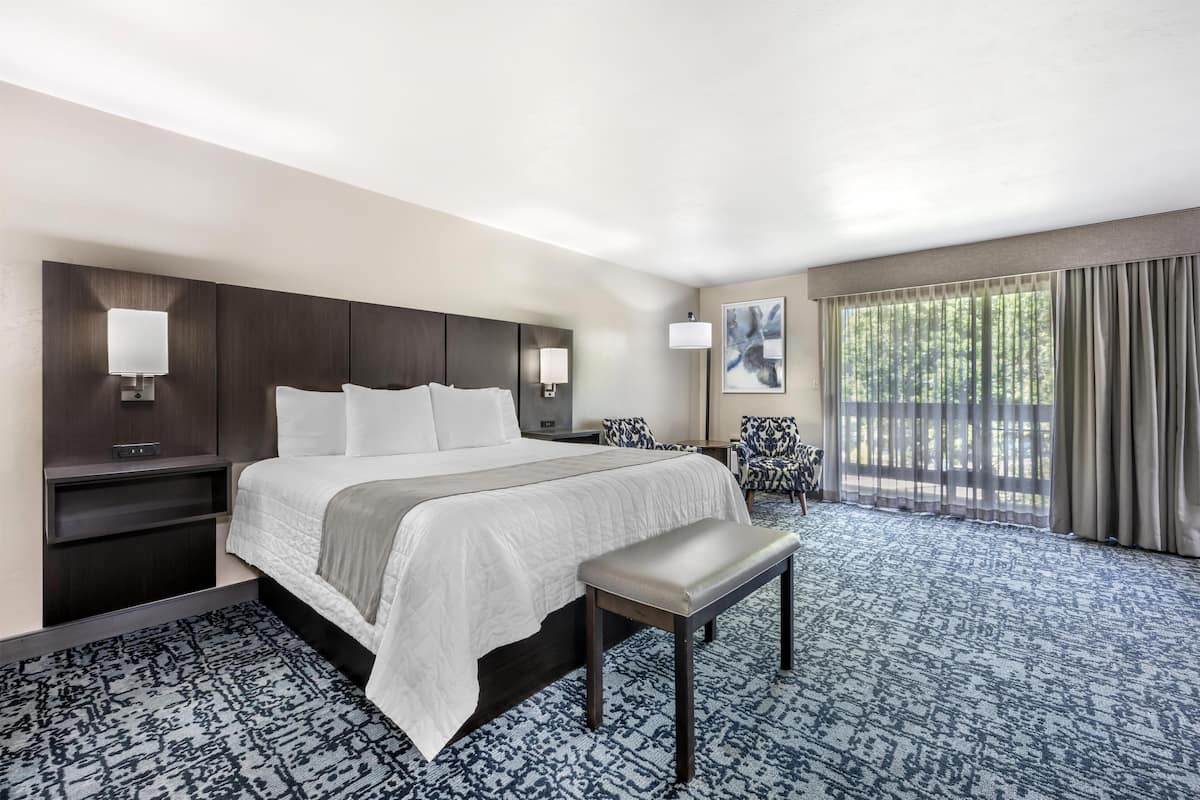 A hotel room with a large bed, bedside tables, a bench, a chair, a window with curtains, and a painting on the wall.