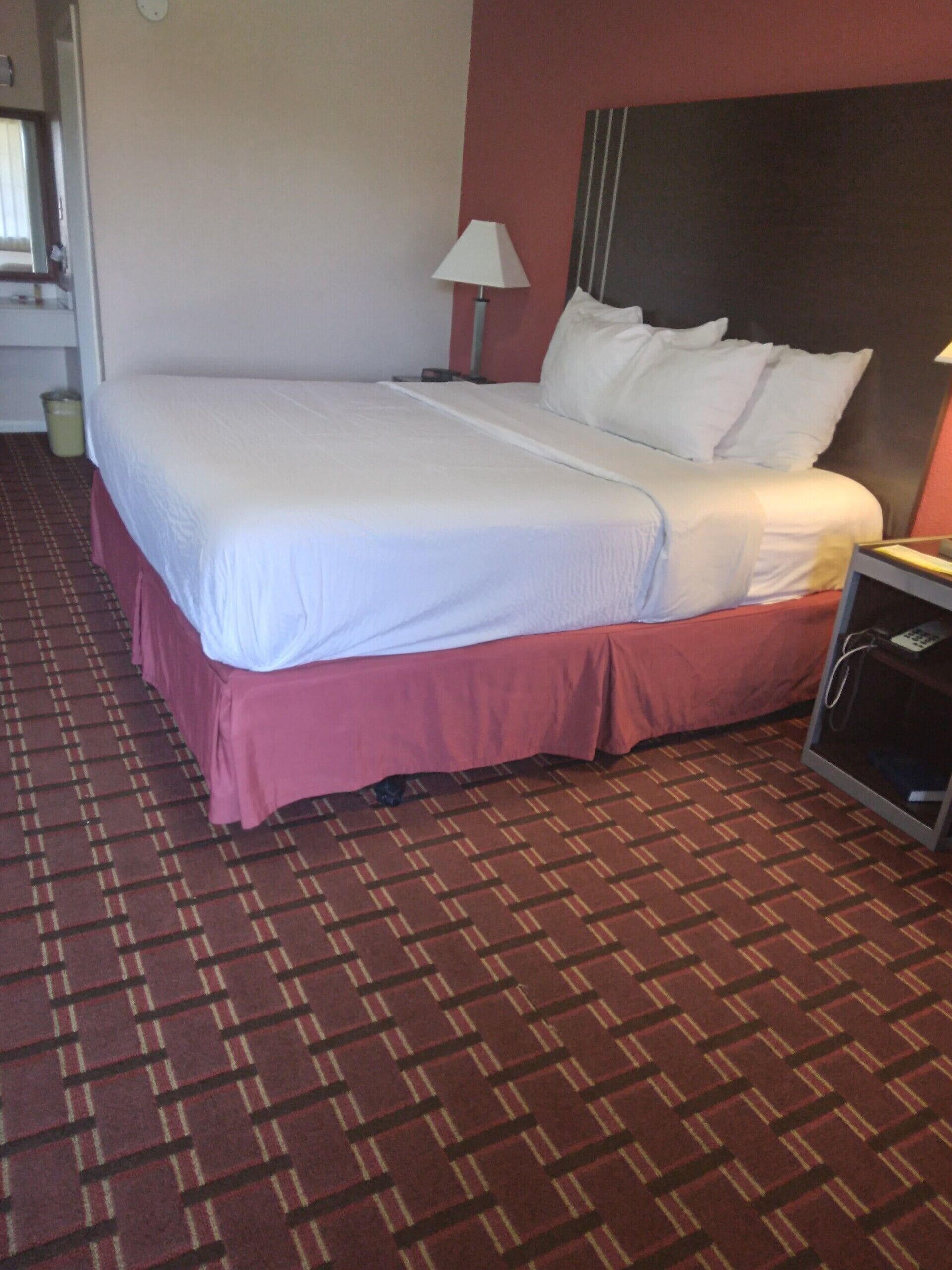 Una habitación de hotel con una cama grande, una mesita de noche con una lámpara y suelo alfombrado.