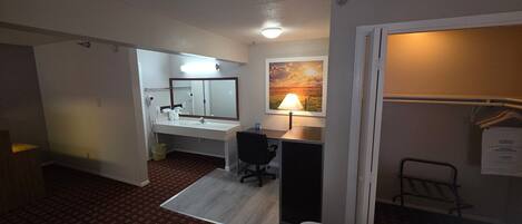 A hotel room with a bathroom, a desk, a chair, a lamp, and a painting on the wall.