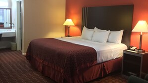 A hotel room with a large bed, two bedside lamps, a nightstand, a desk with a chair, and a bathroom visible through an open door. - Howdy Hotel (College Station)