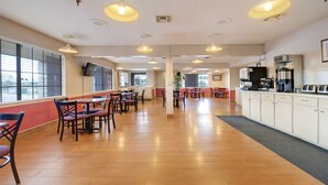 A spacious dining area with multiple tables and chairs, a kitchen area with appliances, and large windows with blinds. - Howdy Hotel (College Station)