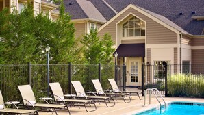 A swimming pool with lounge chairs and a fence, surrounded by greenery and a building with a gabled roof.