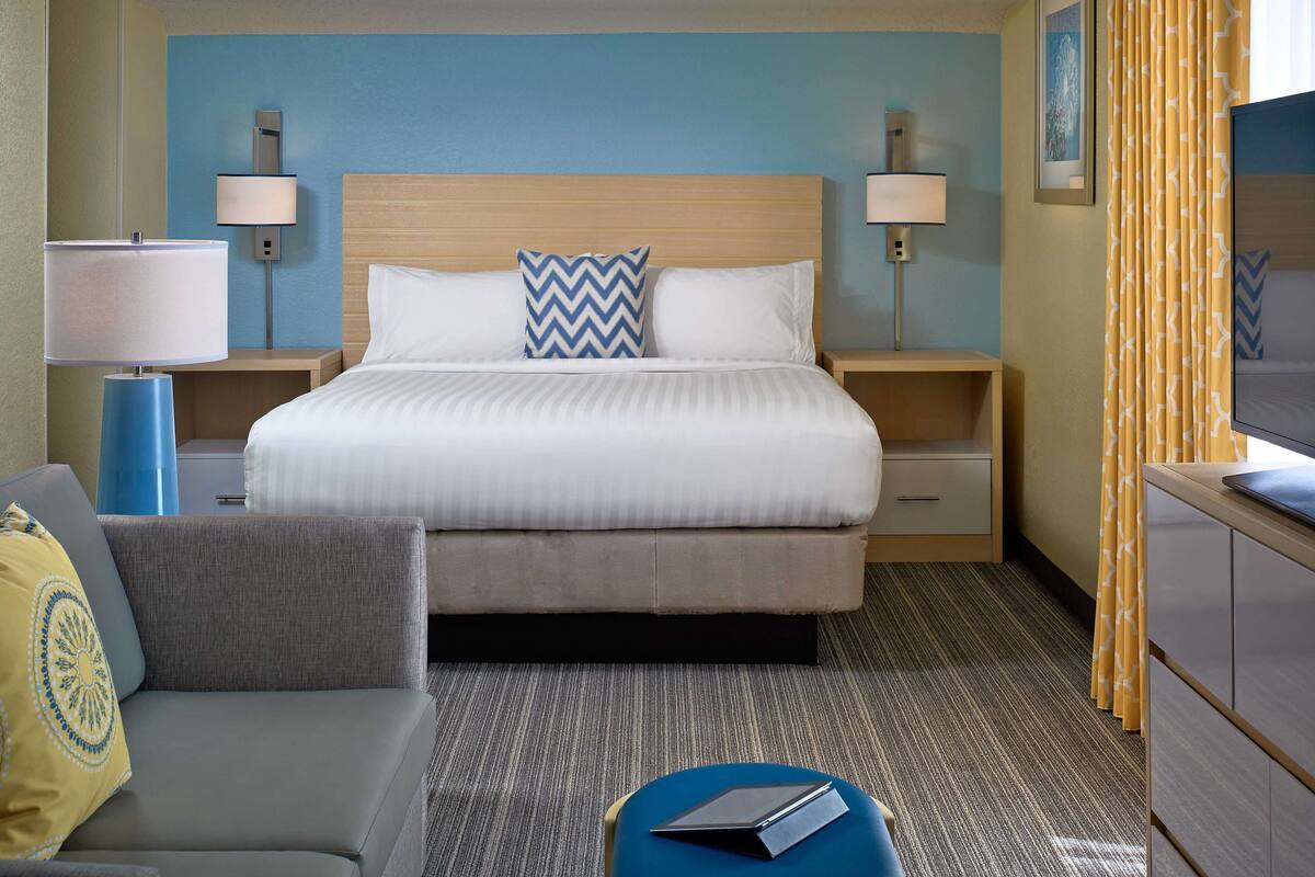 Quarto de hotel com cama, escrivaninha, cadeira, luminária e televisão.