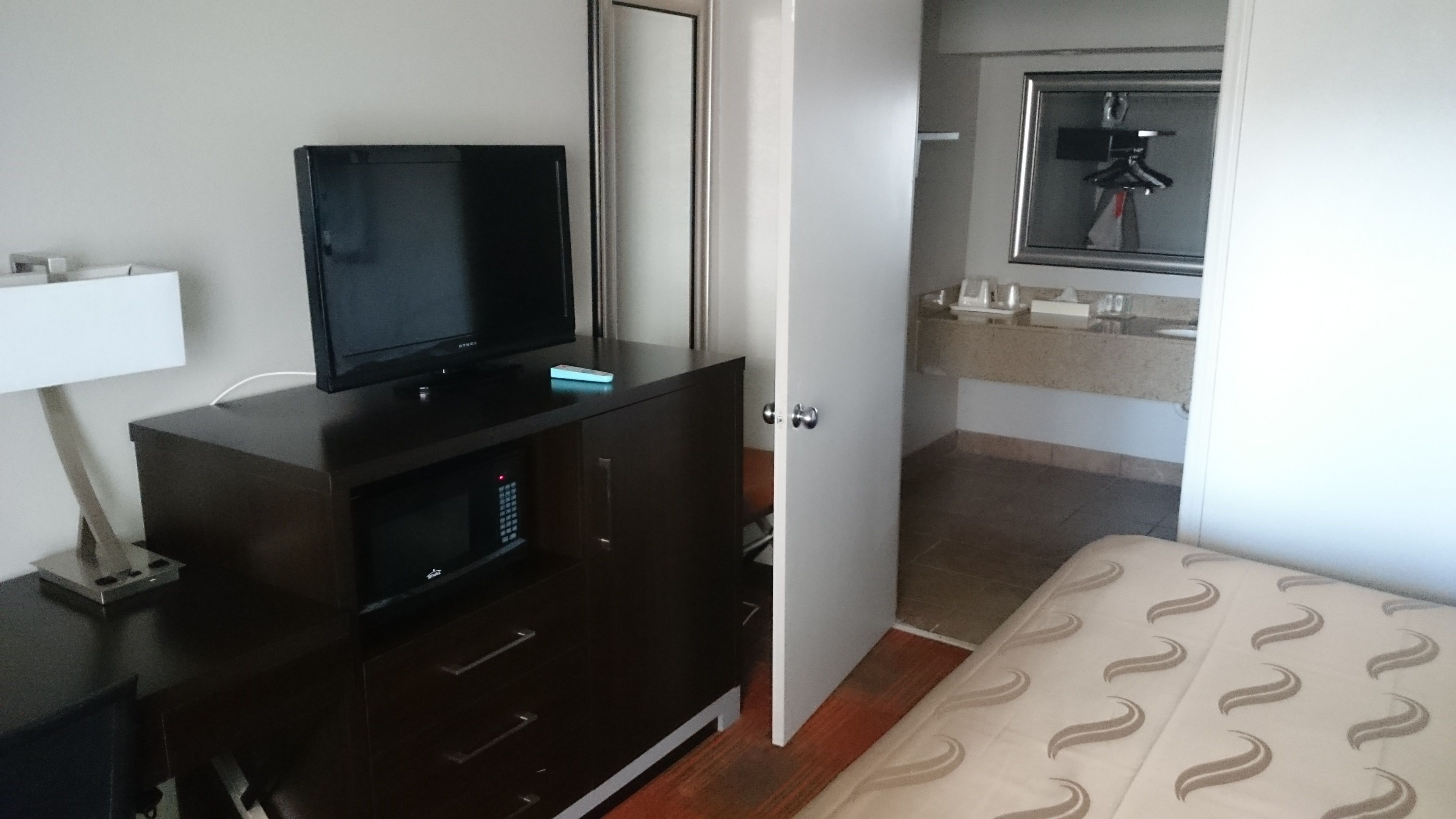 A hotel room with a dark wooden dresser, a flat-screen TV, a bedside table with a lamp, a bed with a patterned bedspread, and a bathroom with a mirror and towel rack.
