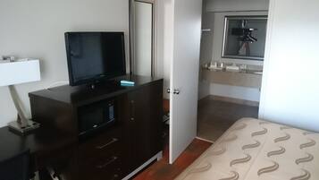 A hotel room with a dark wooden dresser, a flat-screen TV, a bedside table with a lamp, a bed with a patterned bedspread, and a bathroom with a mirror and towel rack.