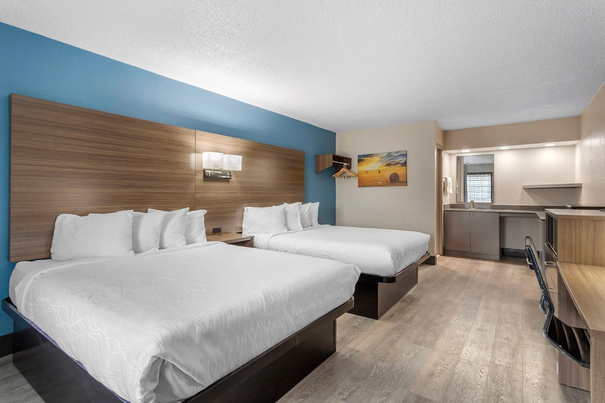 A hotel room with two beds, a kitchenette, and a painting on the wall.
