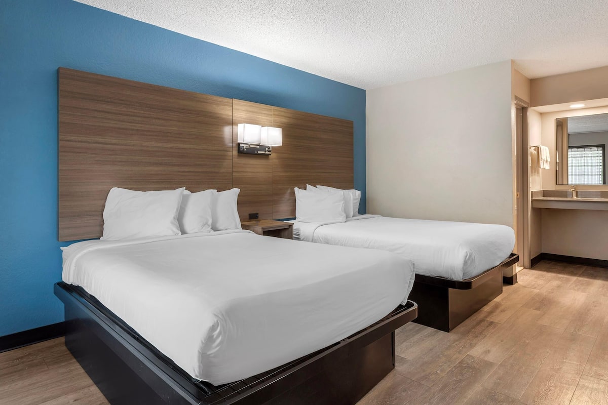 A hotel room with two beds, a wooden headboard, a nightstand, and a bathroom with a mirror and sink.