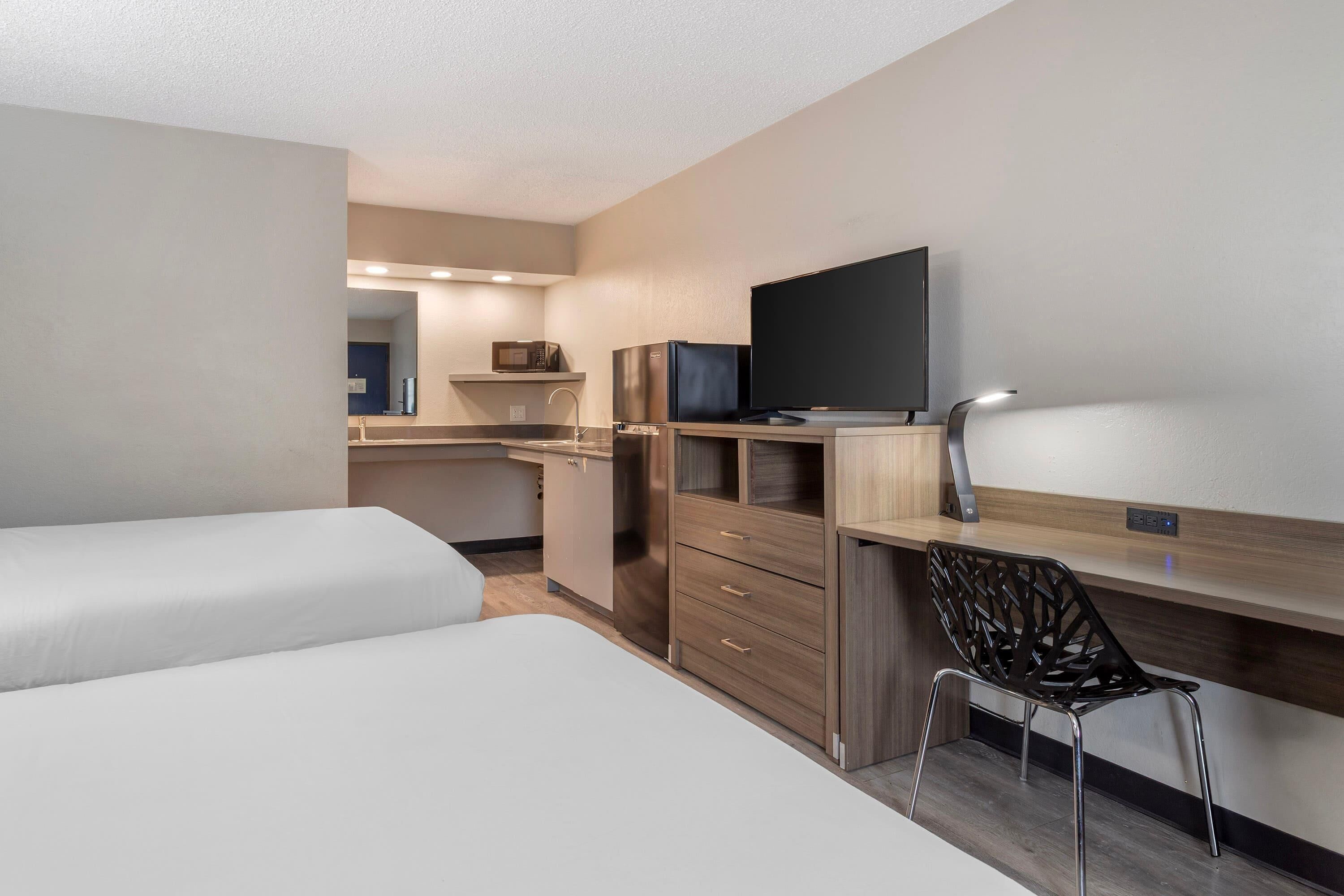 A compact hotel room with a kitchenette, a bed, a desk with a chair, and a TV.