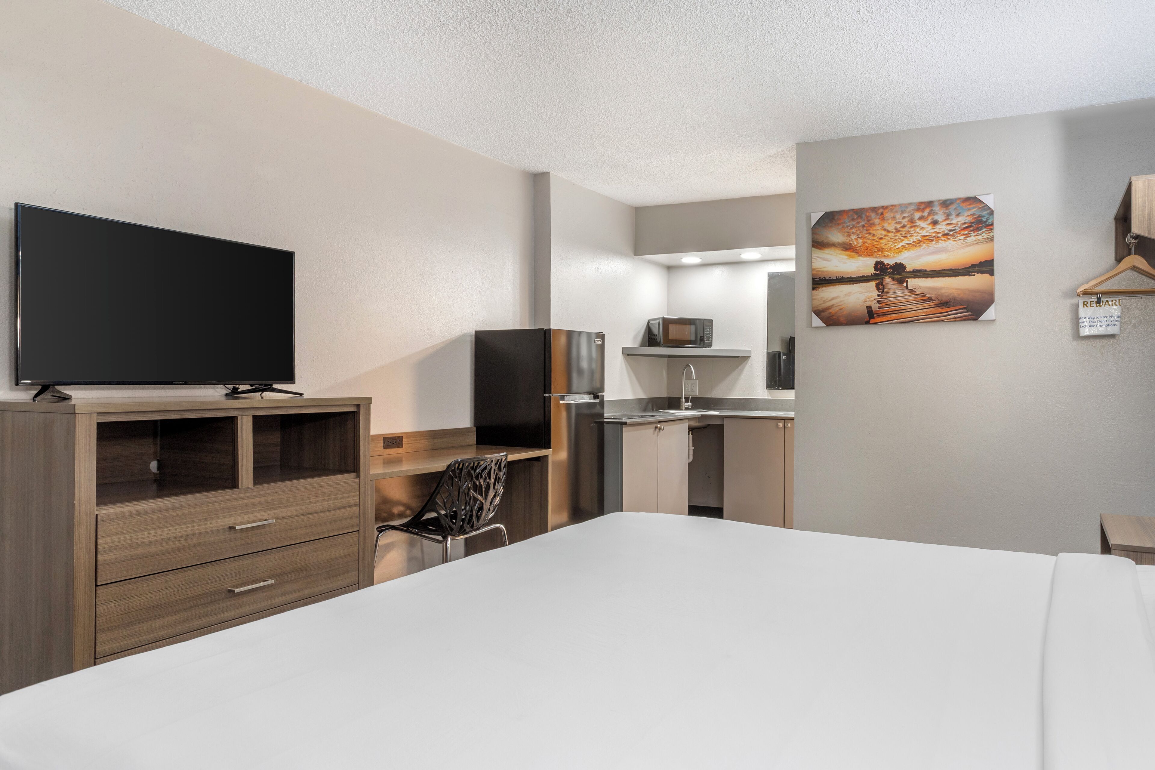 A hotel room with a bed, a TV, a kitchenette, and a painting on the wall.